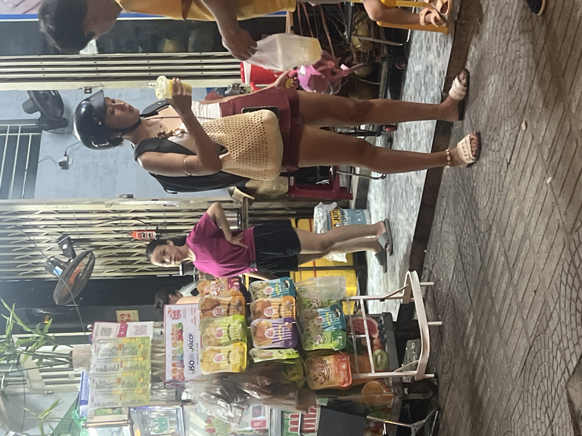 The image depicts two women on a street in Da Nang, Vietnam, likely at dusk or night, given the artificial lighting. The setting appears to be a bustling street or market area.
In the foreground, a woman wearing a pink t-shirt and black shorts stands with her hands on her hips, looking towards the right. She is wearing grey slides. Behind her, to the left, is a display of packaged snacks, possibly in a market stall. Various boxes and bags of food items are visible, with some featuring colorful packaging and possibly text in Vietnamese or other Asian languages.
In the background, another woman is seen wearing a black top, a white undergarment, and maroon shorts. She has a black helmet on and is holding a yellow beverage cup to her lips. She is also wearing white sandals and a woven bag is slung over her shoulder. To the left of this woman, a black fan is mounted on a wall. A glimpse of another person with a yellow shirt is visible in the top left corner.
The ground is paved with patterned tiles. The overall impression is a snapshot of everyday life on a street in Vietnam, with commercial activity and people going about their routines. Some visible text includes "ISO HACCР" on a box of food products. The lighting suggests it is not daytime, possibly evening or night.