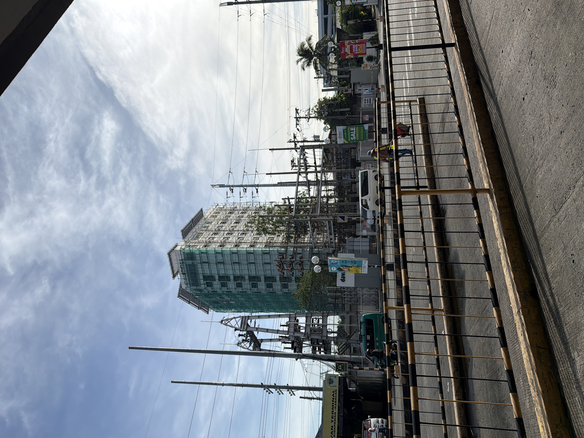 An eye-level, medium-shot photograph depicts an urban street scene in Davao City, Philippines. The image is oriented sideways, with the sky on the left and the ground on the right.
A tall building under construction is the focal point. It is covered in green scaffolding and protective netting. Power lines and utility poles crisscross the foreground. A yellow and black metal barrier runs along the edge of the road. A white van and a green jeepney are parked near the barrier. Two individuals wearing safety vests are visible on the sidewalk.
In the background, there are other buildings, trees, and advertisements. A sign indicating "VAN TERMINAL KIDAPAWAN-KABACAN" is visible. The sky is partly cloudy.