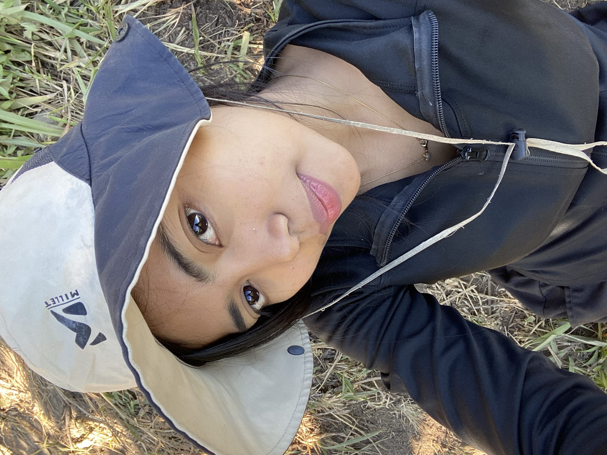 A young Indonesian woman, seen from a high-angle perspective, is lying on dry grass and possibly dirt, with some green vegetation scattered around. She is wearing a wide-brimmed hat, dark jacket, and dark pants. The hat is light-colored with a dark brim and features the "Millet" logo. She has dark hair and is looking towards the camera with a slight smile. A thin, light-colored cord runs from the bottom of the frame up to the zipper of her jacket, and a thin gold necklace with a small pendant is visible. The lighting suggests it is daytime. The scene appears to be outdoors in a natural, undeveloped area.