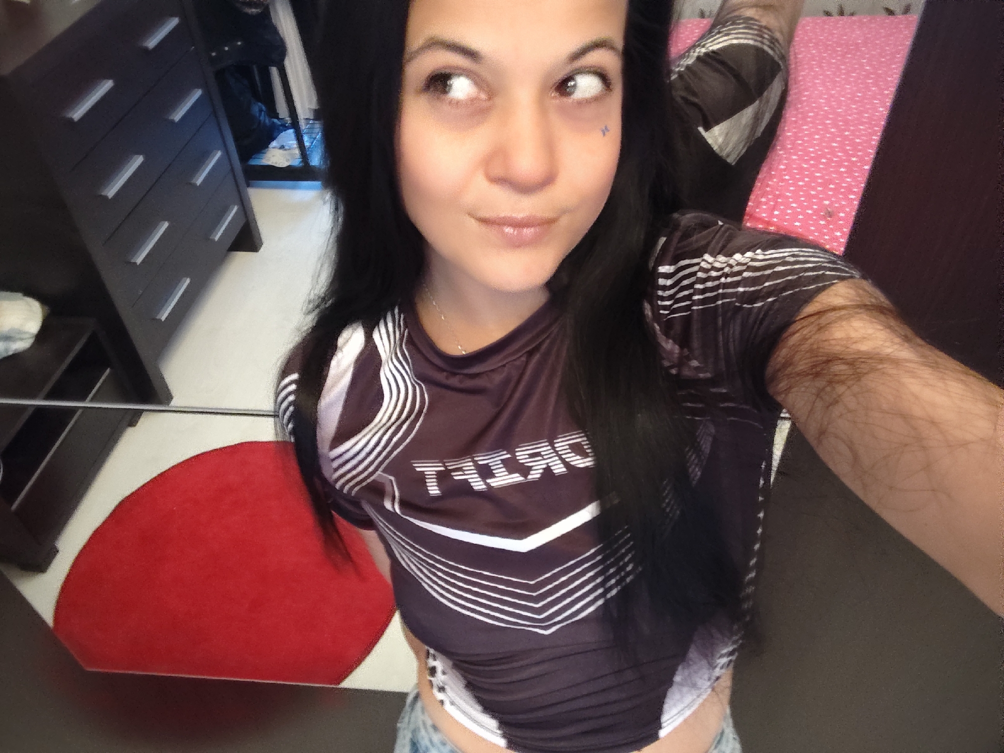 A woman is taking a selfie in a room. She has long dark hair and is wearing a dark t-shirt with white stripes. Her right arm is resting on her leg, which is covered in hair. In the background, there is a dresser with several drawers, a red circular rug, and a bed with a pink patterned sheet. The overall environment appears to be a bedroom or living space. The location is unknown.