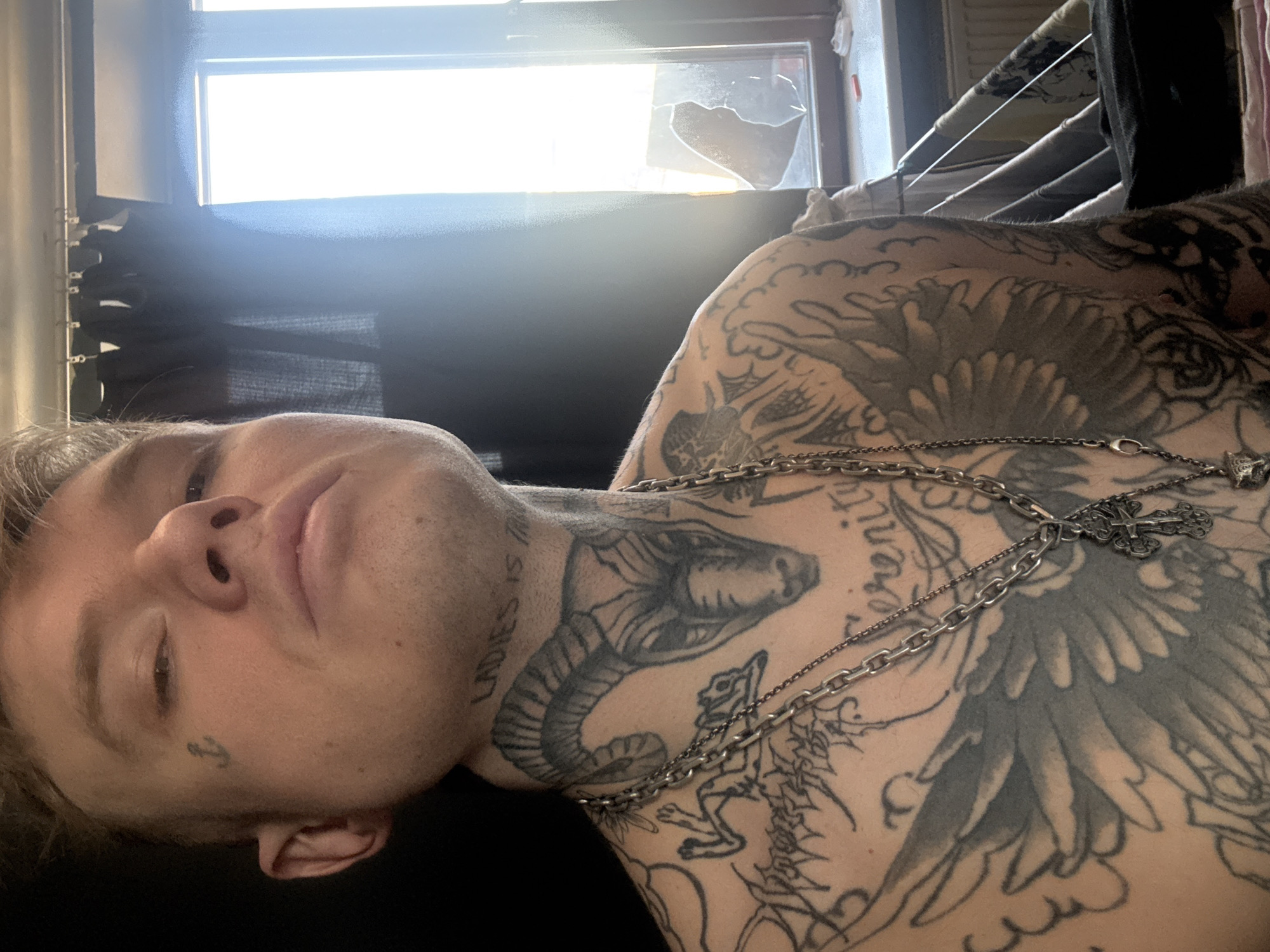 A man with numerous tattoos is lying down. The tattoos cover his neck, chest, and upper arms, including a cobra on his neck, a wing design on his chest, and text on the side of his neck that reads "LADIES IS TH..." (the word is cut off). He is wearing two silver chains with a cross pendant. He has short blonde hair, and a small anchor tattoo is visible near his left eye.
In the background is a window with a broken pane of glass, a dark curtain, and what appears to be a clothes drying rack.
No specific location in Moscow, Russia can be discerned from this image.