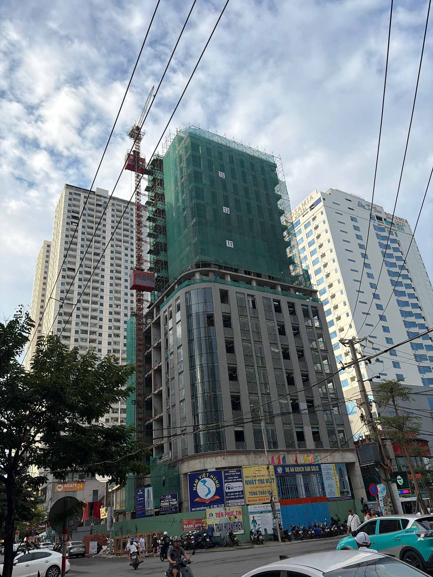 The image shows a street scene in Da Nang, Vietnam, with several tall buildings under construction. A red construction crane is visible, with a green net draped over a partially built skyscraper. Adjacent to the construction site, a completed white skyscraper with blue-tinted windows stands tall. The street below is busy with traffic, including motorcycles, cars, and pedestrians. Billboards and signs with Vietnamese text are displayed on the buildings. The sky is partially cloudy, indicating daylight hours. The overall scene suggests an urban environment experiencing ongoing development.