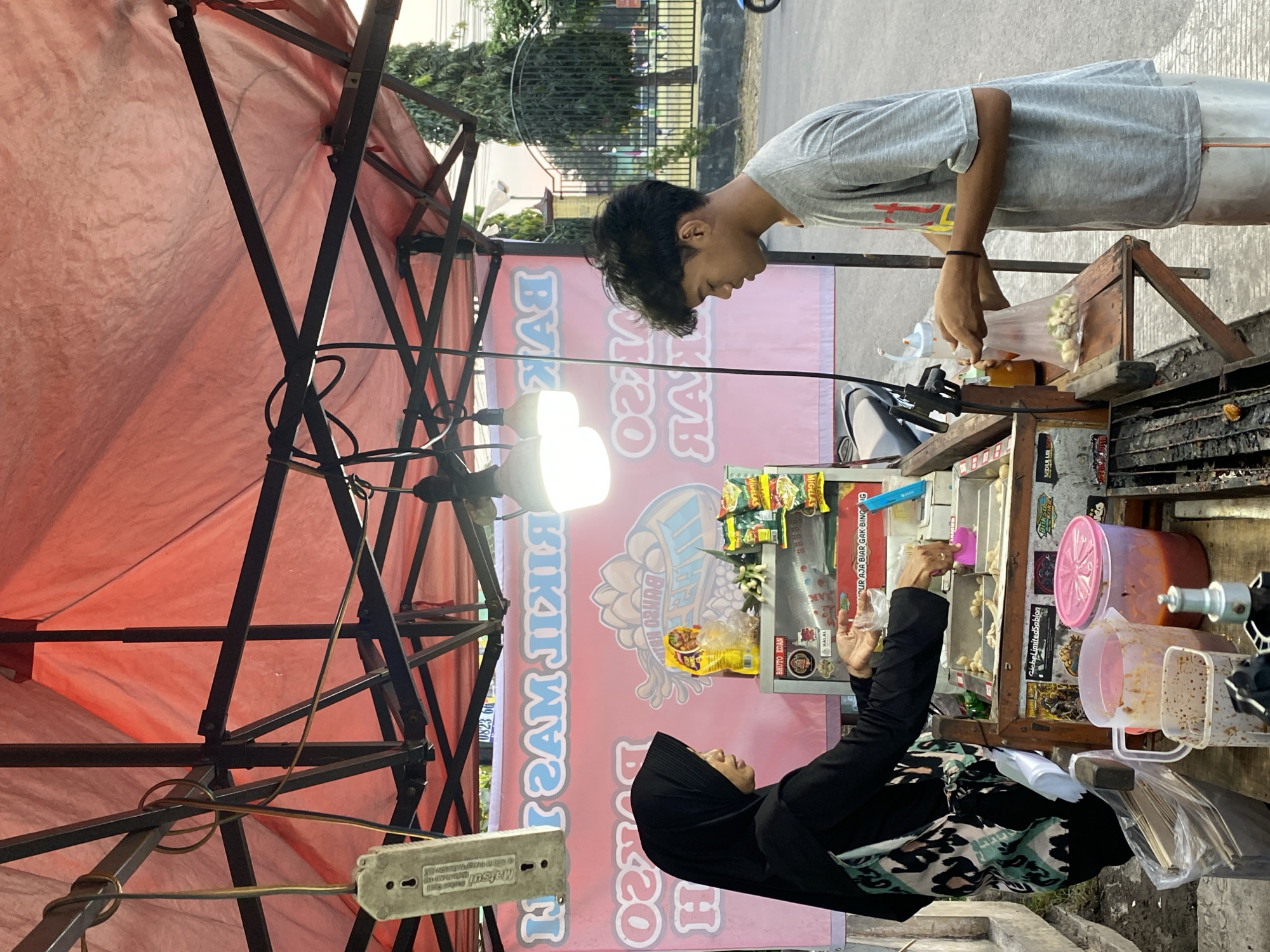 The image depicts an outdoor food stall at street level, likely in Kasihan, Indonesia, as suggested by the context. A woman wearing a black hijab and a patterned vest is serving a customer, a young man in a grey t-shirt. The stall is covered by a red tarp supported by a metal frame, with several light bulbs illuminating the scene. Behind the stall, a pink banner with Indonesian text, possibly advertising "Bakso" (meatball soup), is visible. The woman is holding a plastic bag, appearing to be preparing an order for the young man, who is also holding a plastic bag. Various food items, containers of sauce, and packaging materials are arranged on the stall's counter. The background shows a street, a metal fence, trees, and some distant buildings, suggesting an urban or suburban environment. The overall atmosphere is that of a casual, everyday transaction at a local food vendor.