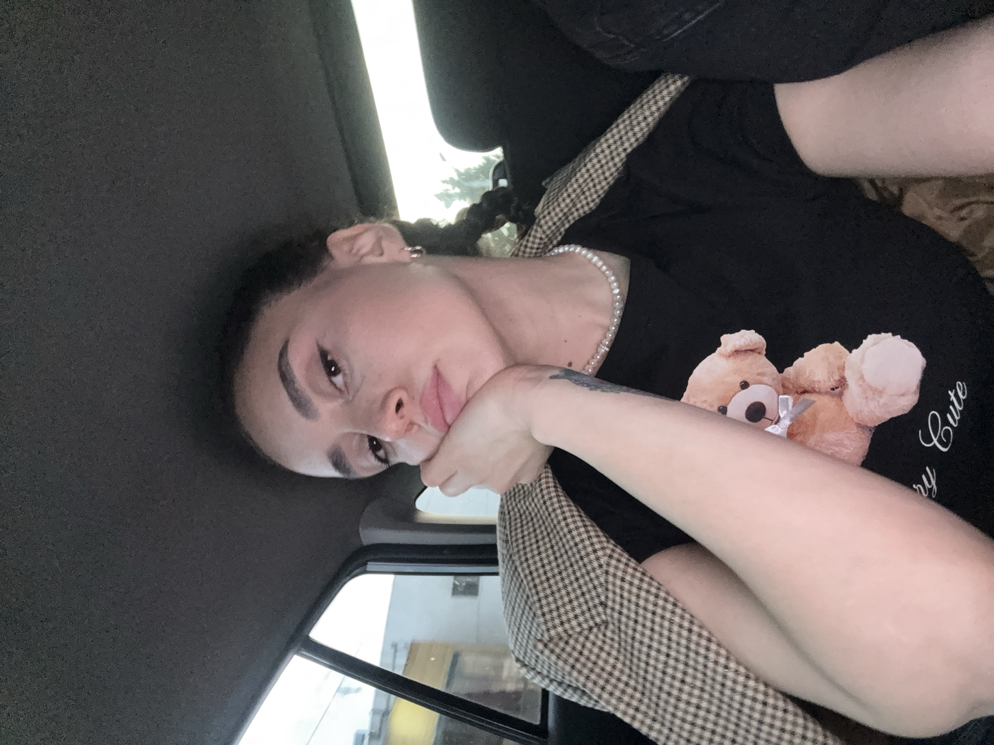 A young adult is seated in what appears to be the rear of a vehicle. The subject has light skin, braided hair, dark eyebrows, and a pearl necklace. They are wearing a black t-shirt with a teddy bear graphic and "very cute" text. A seatbelt with a brown and white houndstooth pattern is visible. The subject's left hand is touching their face. The vehicle's interior is predominantly dark gray. Through the car window, indistinct buildings and daylight suggest an urban environment; based on the prompt, this would be in Tbilisi, Georgia.
