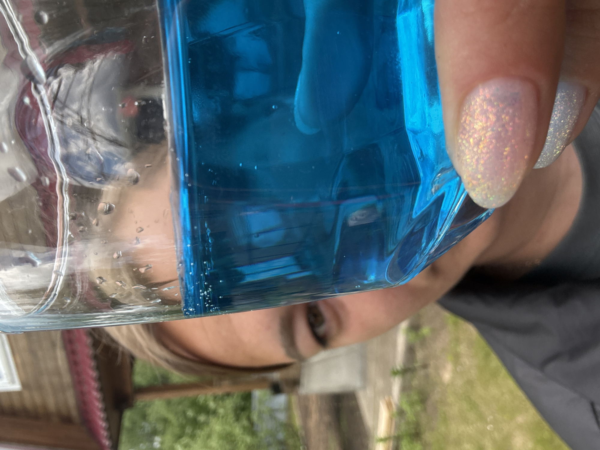 The image is an extreme close-up perspective, likely a selfie, of a person holding a clear and blue liquid-filled container, presumably a bottle. The person's face is visible behind and distorted by the bottle, with one eye clearly visible. Their hand and fingernails are also in view, the nails painted with a light iridescent glitter polish. The background is out of focus and depicts an outdoor scene with greenery, a building with a visible window frame and wooden structure, potentially indicating the city of Dmitrov, Russia, based on implied architectural styles commonly found there, and partial view of a dark gray or black colored garment. The scene suggests an ordinary, casual moment of interaction with objects in an outdoor environment.