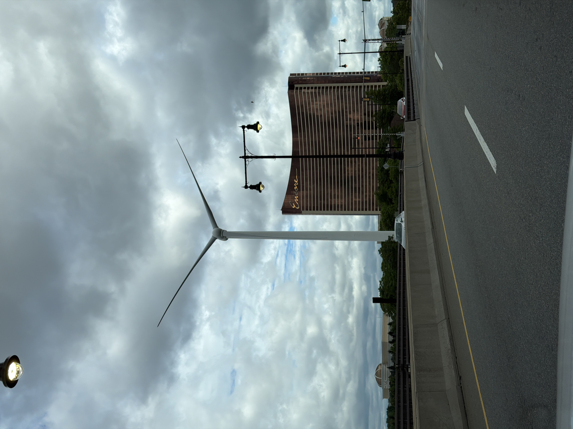 The image depicts a cloudy day view from a highway in Everett, Massachusetts, part of the greater Boston area. In the foreground, an asphalt highway with white lane markings curves to the right. A concrete barrier separates the highway from a raised area with some trees and a dark structure. In the mid-ground, a large, modern building with a copper-colored facade and the word "Encore" in gold lettering is visible. To the left of the Encore building, a tall, white wind turbine stands prominently, its blades angled upward. Two ornate streetlights with illuminated bulbs are positioned in front of the Encore building. In the background, more trees and buildings, including one with a domed roof, can be seen under the overcast sky. A silver car is parked on the raised area, and another red car is visible further down the road. There are no people visible in the image.
