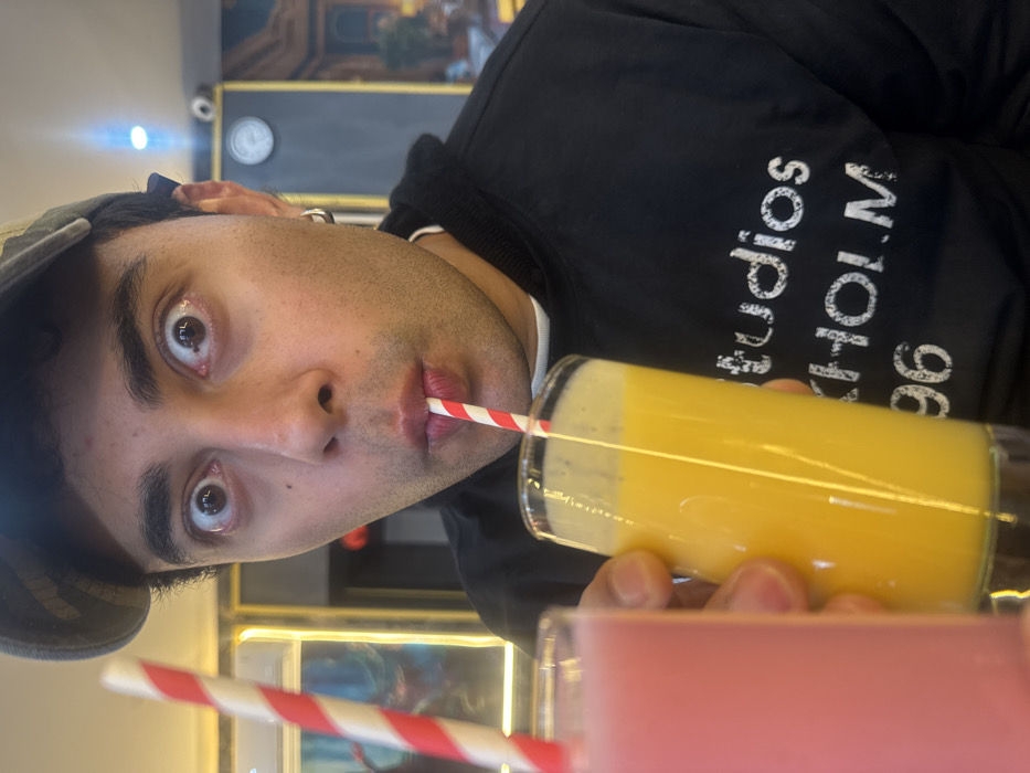 A male individual is captured in a close-up, eye-level shot within an indoor commercial establishment in Paris, France. He wears a camouflage-patterned baseball cap and a black hooded sweatshirt with partial white text "studios STKHOLM 96" visible on the chest. A silver hoop earring is present in his left ear. He is actively drinking from a tall glass containing a bright yellow/orange beverage, using a red and white striped straw. His eyes are wide and directed towards the camera, while his mouth is pursed around the straw. His right hand holds the glass. To his left, another tall glass containing a pink beverage, also with a red and white striped straw, is partially visible and out of focus. The blurred background reveals elements consistent with a cafe or juice bar, including a light-colored wall, a gold-framed object (possibly a mirror or artwork), a darker wall section, a white circular fixture, and a security camera (CCTV) mounted high in the upper right. A distinct blue light source is also visible in the upper background.