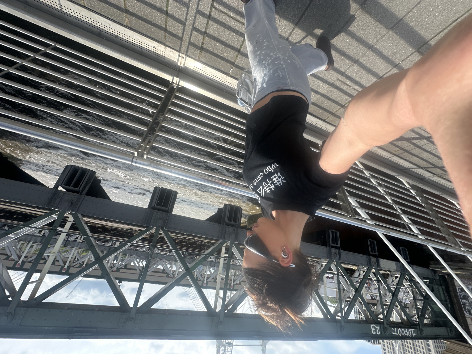 A person is standing on a bridge in London, United Kingdom, taking a selfie. The photo is rotated such that the person appears upside down. The bridge structure is visible below, with the river running alongside. The person is wearing a black t-shirt, light-colored jeans, and sunglasses, and has stretched ear piercings. The bridge has metal railings, and the walkway is paved. The bridge's structural supports and the river are visible in the background. The bridge is labeled with the words "FOOT 23".
