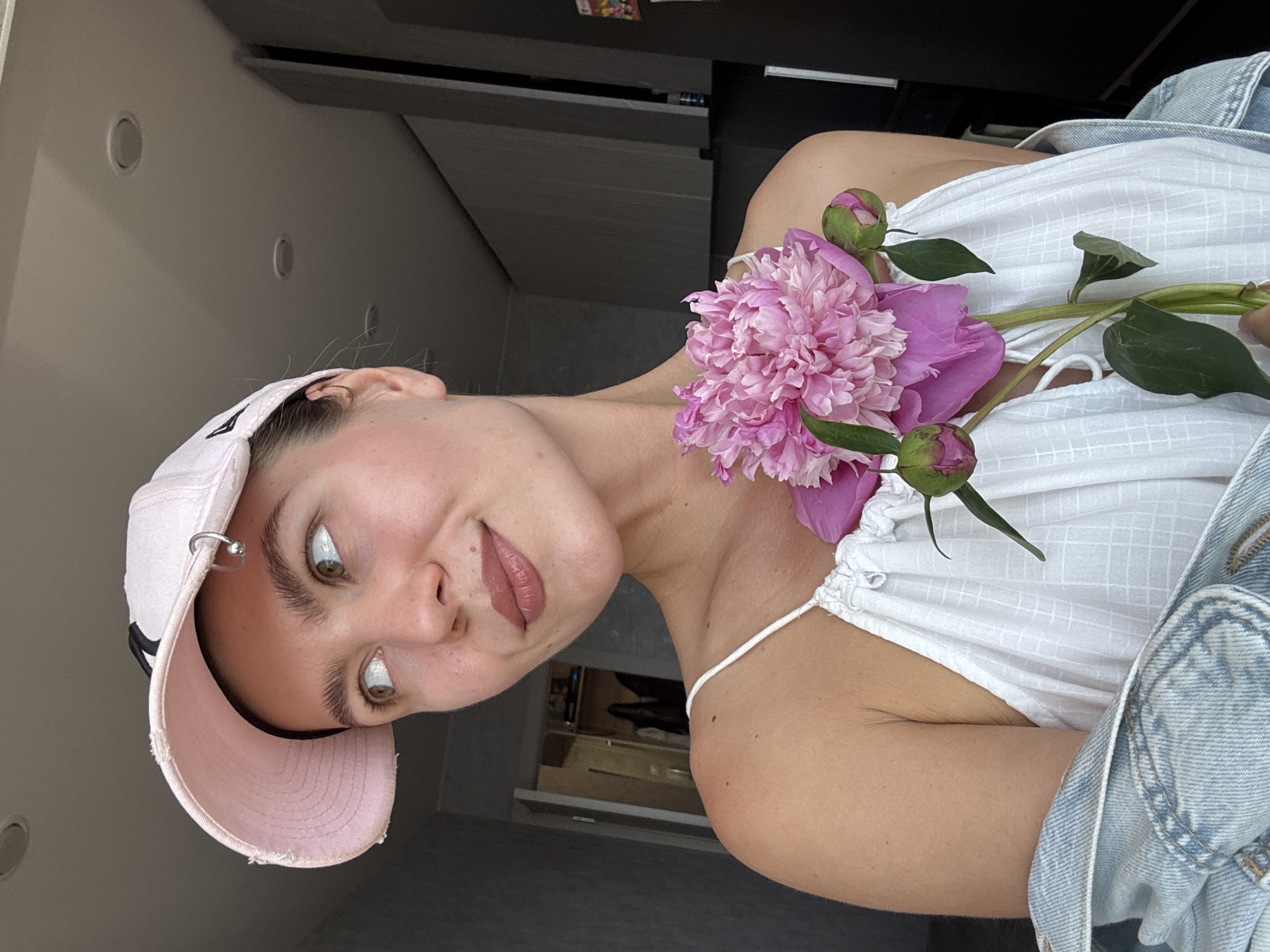 A young woman with fair skin, brown eyes, and light brown hair is pictured in a close-up shot, holding a pink peony. She is wearing a pink baseball cap and a white tank top, with a denim jacket draped over her shoulders. She is looking to her left with a slight smile. The background appears to be an indoor environment, possibly a kitchen or living room, with cabinets and lighting fixtures visible. The lighting is soft and natural.