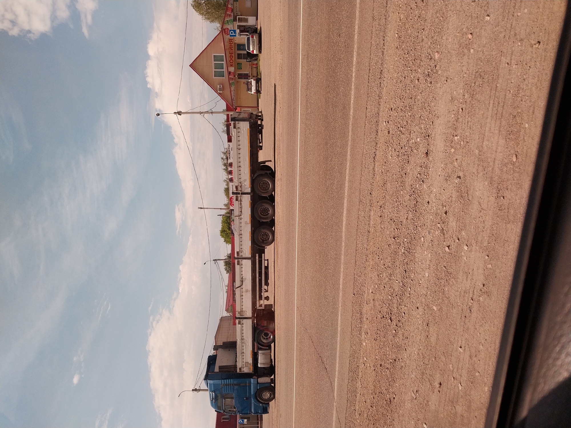 A blue truck with a trailer is parked on the side of a road. The truck is facing to the left, with its front cab on the left side of the image. The trailer is long and flat, with metal railings along the sides. The truck is a semi-trailer truck, and it appears to be empty. The trailer has "ЭВАКУАТОР" written on its side, which translates to "TOW TRUCK."
In the background, there is a small building with a red roof and beige walls. The building has several windows and a door. There is also a sign above the door that says "КОФЕЙНЯ" which means "COFFEE SHOP." In front of the building, there are two white cars parked.
The road is made of asphalt, and there are white lines painted on it. The scene appears to be a rural or suburban area. The sky is blue with some white clouds. The sun is shining, and it appears to be daytime. The overall mood of the image is peaceful and still.
A power line runs across the top of the image, with a lamppost visible near the truck. The image is taken from a slightly elevated angle, looking down at the truck and the building. The quality of the image is clear, and the colors are vibrant.