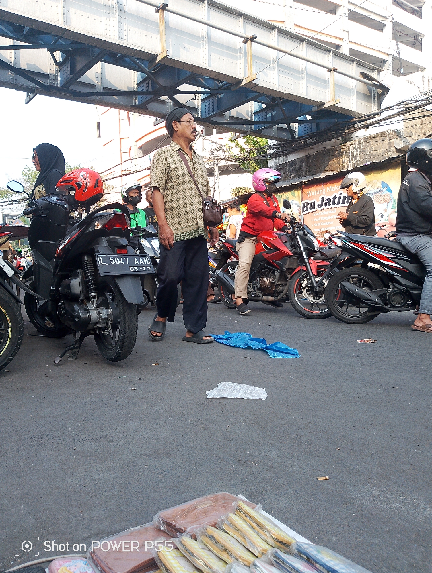 A street scene in Surabaya, Indonesia, shows a group of people and motorcycles gathered under an elevated structure, possibly a bridge or overpass. The foreground features a black motorcycle with a license plate reading "L 5047 AAH". A man in a patterned shirt and dark pants is walking towards the viewer, while other individuals, some wearing helmets, are either on or near motorcycles. In the background, a building with windows and wires is visible. The street is paved, and there are some plastic bags and debris scattered on the ground. The lighting suggests it is daytime.
