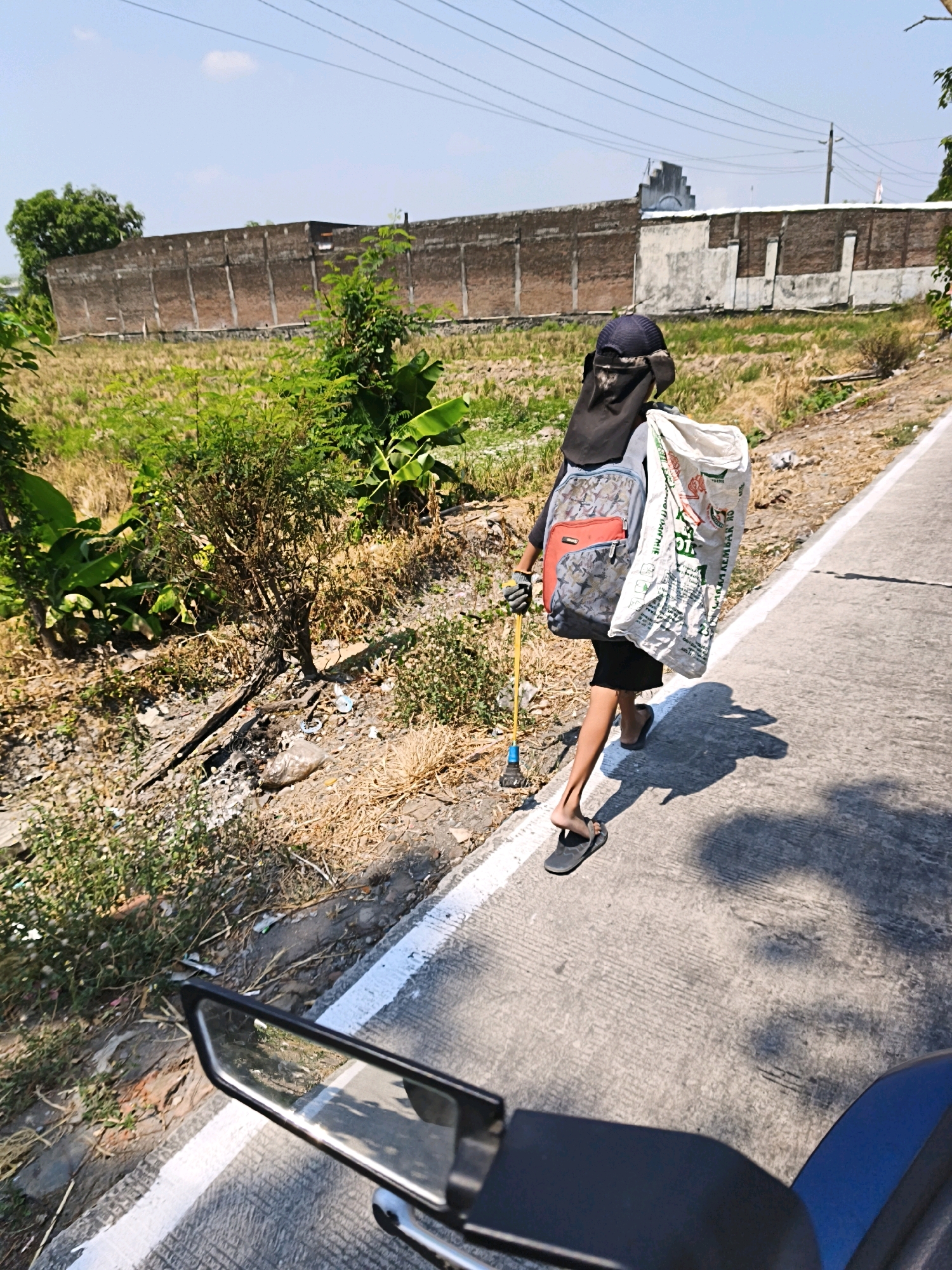 A person wearing a dark head covering, a black shirt, shorts, and flip-flops walks on the side of a concrete road in Desa Donohudan, Indonesia. The individual carries a large white bag on their right shoulder and a backpack with red and gray accents on their left. In their right hand, they hold a yellow stick with a blue object at the end, possibly a litter picker. The person casts a long shadow on the road, indicating a sunny day. To the left of the road is a dry, overgrown area with sparse vegetation, rocks, and scattered debris. A brick wall, partially covered in white plaster, runs along the background, separating the roadside from a field of dry grass and some green plants. A dark blue vehicle part is visible in the lower right corner, suggesting the image was taken from a vehicle. The sky is clear and blue with a few thin clouds.