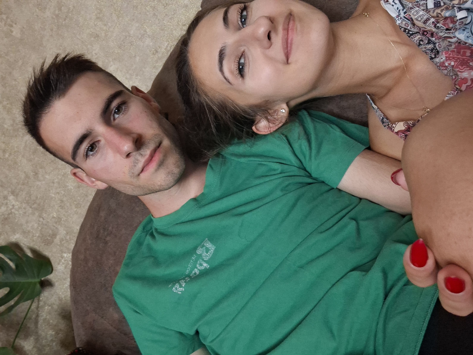 Two people, a man and a woman, are lying down on a brown sofa. The man is on the left, wearing a green t-shirt with a white logo. The woman is on the right, wearing a patterned dress and a gold necklace. She has red nail polish on her fingernails. Both are looking towards the camera and smiling. The background appears to be a wall with a textured pattern. A large green leaf plant is visible in the upper left corner. The lighting is soft, suggesting an indoor setting.