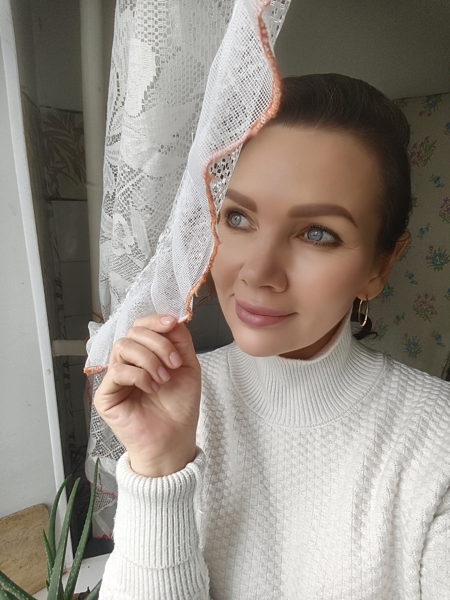 A Caucasian woman is depicted in a medium shot indoors, wearing a white, high-necked sweater and gold hoop earrings. She is holding a white lace curtain with a peach-colored trim, looking off to the side with a slight smile. The room has wallpaper with floral designs and a window with a partially visible plant on the sill. The city of Unknown, Unknown cannot be determined from the image.