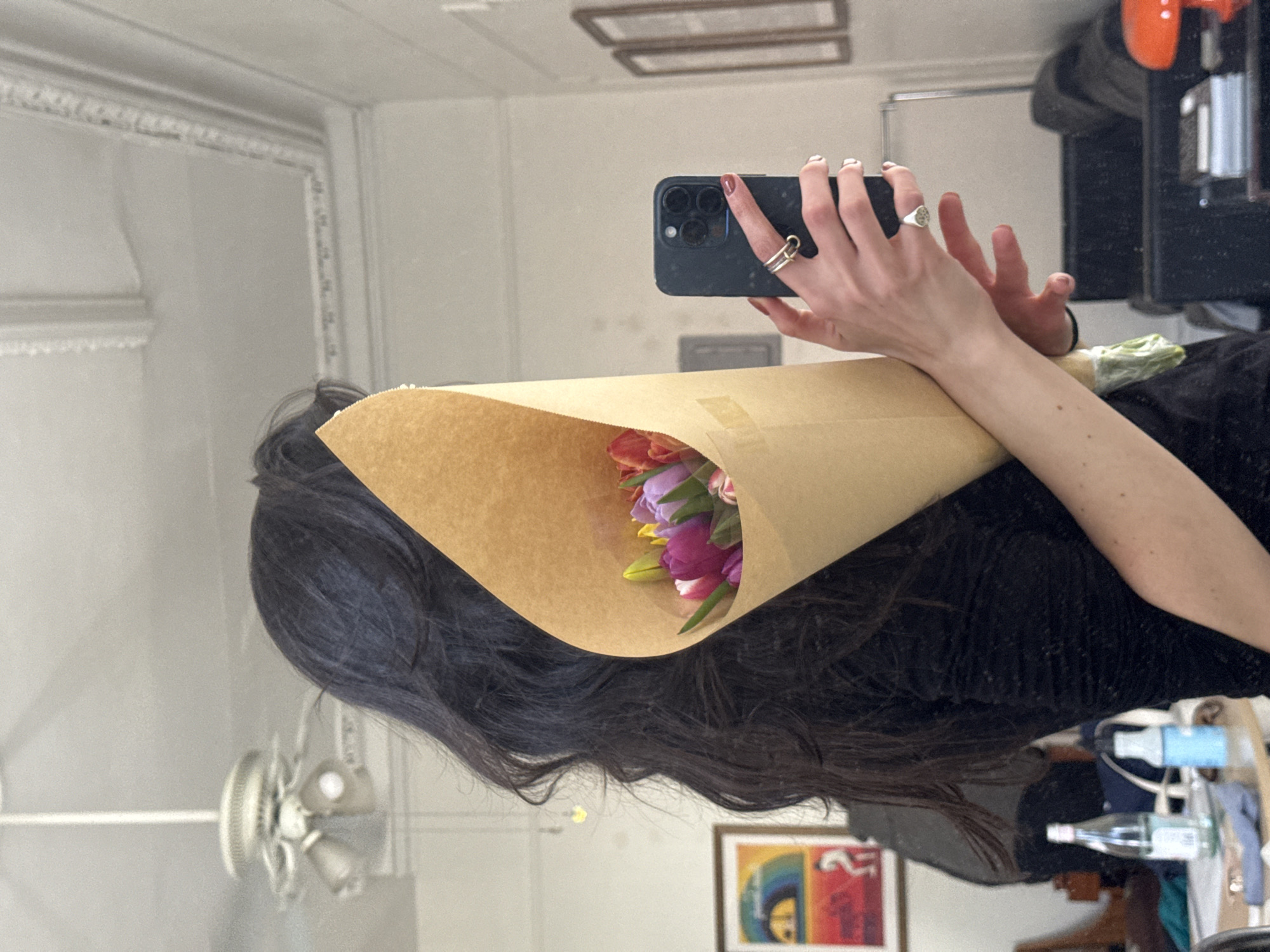 A person with dark hair is holding a bouquet of tulips wrapped in brown paper. They are using a smartphone to take a picture of themselves in a mirror. The reflection in the mirror shows architectural details, a ceiling fan, and a framed picture. In the background, on a table, are water bottles, other objects and a black dresser. The reflection suggests the location is in an apartment or home within an urban setting, likely in New York City, United States of America. The subject is facing away from the viewer.
