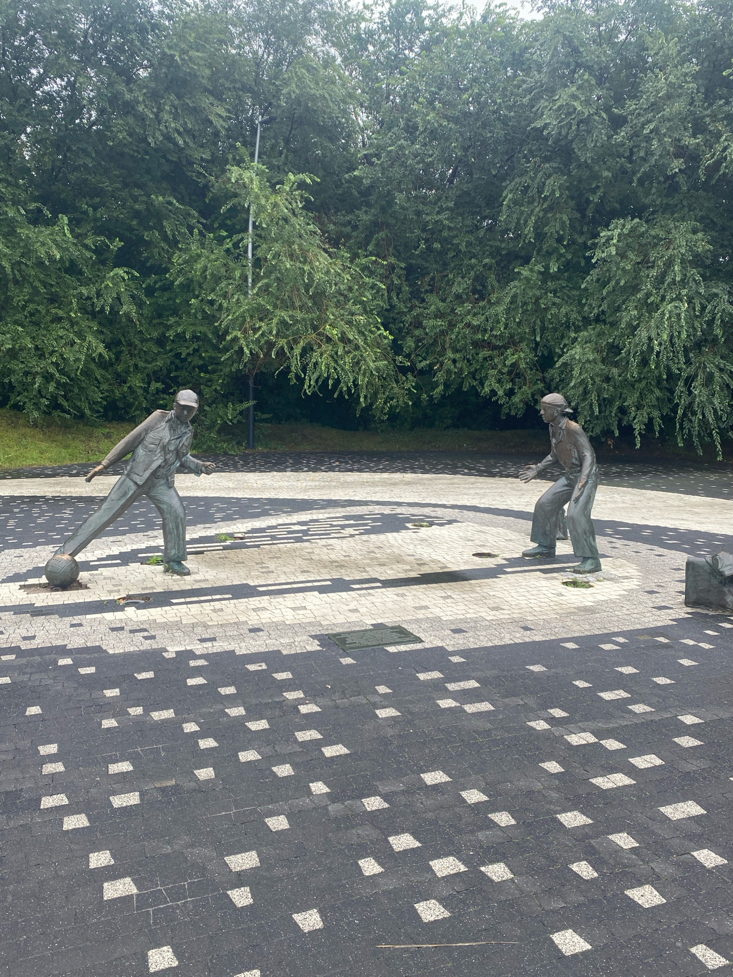The image depicts a bronze sculpture of two men playing football, located in Kazan, Russia. The sculpture is situated on a paved plaza with a distinctive checkered pattern of dark and light square tiles. In the foreground, one man is captured mid-action, kicking a football with his right foot, leaning forward with his arms outstretched. He wears a cap, a jacket, and trousers. The second man, in the middle ground, stands in a defensive stance, facing the other player with his arms held out. He also wears a cap, shirt, and trousers. Behind the statues, a dense line of green trees forms a natural backdrop. The lighting suggests it is daytime, with shadows cast on the plaza, indicating sunlight.