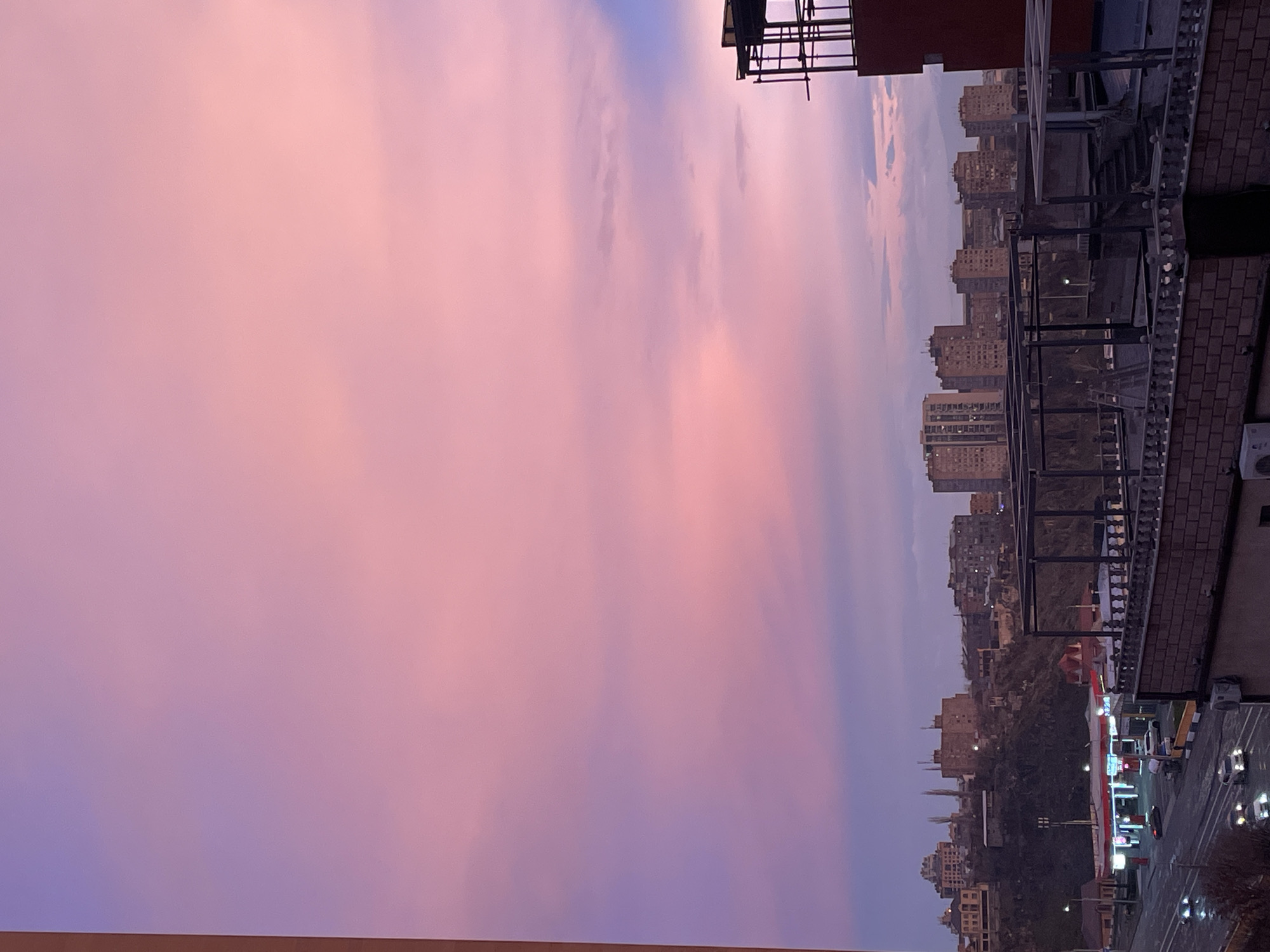The image depicts an urban landscape in Yerevan, Armenia, captured at twilight. The upper two-thirds of the scene are dominated by a vibrant sky exhibiting strong gradients of pink and purple hues, with subtle horizontal cloud formations.
In the immediate foreground, on the right side, a partial view of a building's upper section is visible, featuring dark, open-frame architectural elements or scaffolding against a reddish-orange wall. A brick railing with balusters extends horizontally across the lower portion of the image, below which an external air conditioning unit is mounted on a building wall.
The midground, on the left, features a multi-lane asphalt road with multiple vehicles in motion, identifiable by their illuminated headlights and taillights. Adjacent to the road, a "GP" branded gas station is prominent, distinguished by its red and white structure and illuminated canopy. Directly behind the road and gas station, a dark, moderately sloped hill rises, populated with numerous multi-story residential buildings, some with illuminated windows.
The background extends into a distant cityscape, showcasing a panorama of additional multi-story urban buildings, primarily residential blocks, silhouetted against the colorful horizon. No individual persons are clearly discernible, and observed actions are limited to vehicular movement on the road and at the gas station.