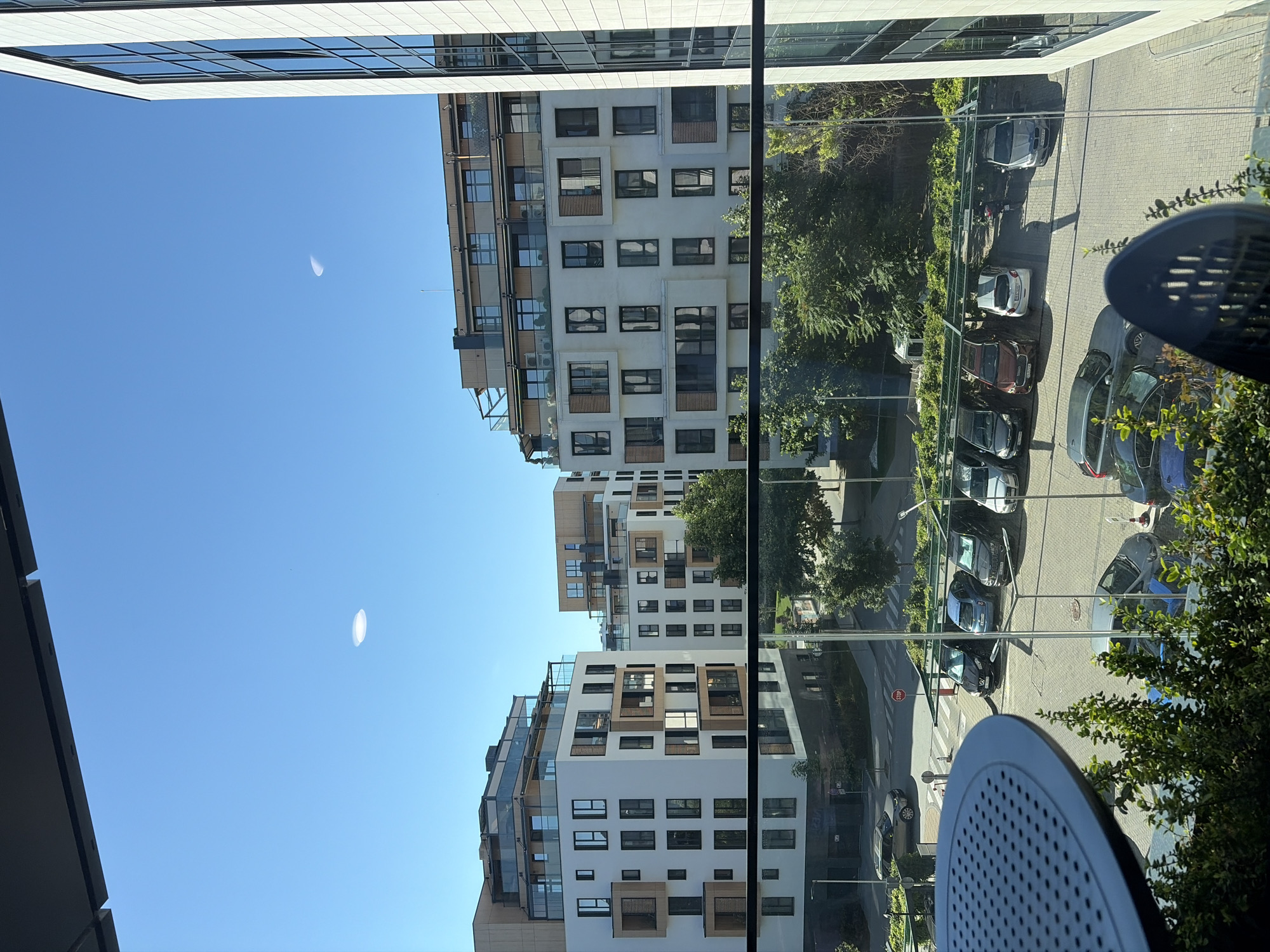 A high-angle view captures a modern city scene with apartment buildings, a parking lot, and vehicles under a clear blue sky. The foreground features a dark circular table with a perforated surface, partially obscuring a lush green bush. Beyond this, a glass railing overlooks a paved area with a parking lot. Several cars are parked, including a silver SUV, a black sedan, a dark blue hatchback, a dark red SUV, a light silver hatchback, and a silver sedan. A motorcycle is also visible in the parking lot. In the background, two contemporary apartment buildings with white facades, dark windows, and wooden accents stand tall. The building on the left has multiple balconies and glass enclosures on its rooftop. The building on the right is partially visible, featuring a modern facade with large windows. A tree-lined area with green foliage separates the apartment buildings and the parking lot. The sky is a bright, cloudless blue with two indistinct oval-shaped light reflections visible.