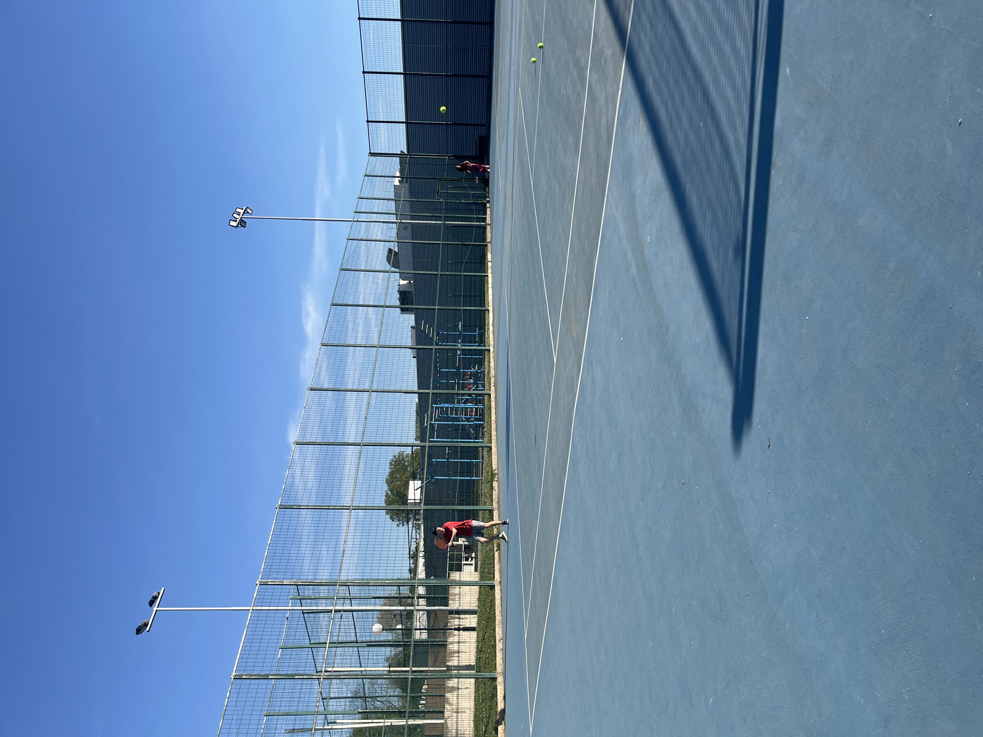 The image displays an outdoor tennis court scene on a bright, sunny day. The photograph appears to be a landscape shot that has been rotated 90 degrees clockwise.
The primary subject is a person, likely male, positioned on the blue-colored hard-surface tennis court. This individual is wearing a red t-shirt, dark shorts, and sneakers, and is holding a tennis racket, appearing to be in the midst of a swing or preparing to hit a ball. Another less distinct figure in a dark shirt is visible further down the court, near the back fence. Three bright yellow tennis balls are scattered across the court surface.
The setting is a tennis court with clear white boundary lines. A tall chain-link fence encloses the playing area, with trees and buildings visible beyond it in the background. The sky above is a clear, vibrant blue with a few faint white clouds. Two tall light poles, each fitted with multiple luminaires, are prominent against the sky, indicating the court is equipped for night play.
The activity taking place is clearly a game or practice of tennis. The strong sunlight casts sharp, distinct shadows from the fence and the light poles onto the court, suggesting it is mid-day or afternoon. There is no visible text in the image.