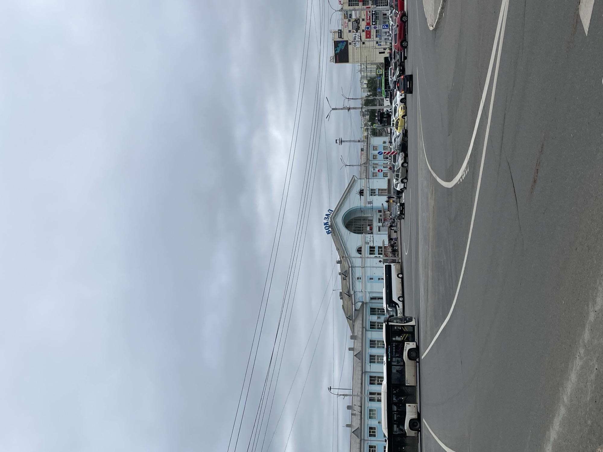 This is a wide shot of a city street scene in Kirov, Russia, on a cloudy day. In the background is a light blue train station building with a large archway and the word "ВОКЗАЛ" (VOKZAL, meaning "station") written in blue lettering on its facade. In front of the station, several cars and buses are visible, suggesting a busy transportation hub. A white bus with an open door is in the foreground on the left, and another white bus is parked near the station. The street is paved and has white lane markings. Power lines stretch across the sky. To the right of the train station, a tall, multi-story building with various advertisements is visible. A traffic light is also present. There are several people walking on the sidewalk near the station and entering/exiting buildings. The overall impression is a functional urban environment centered around a train station.