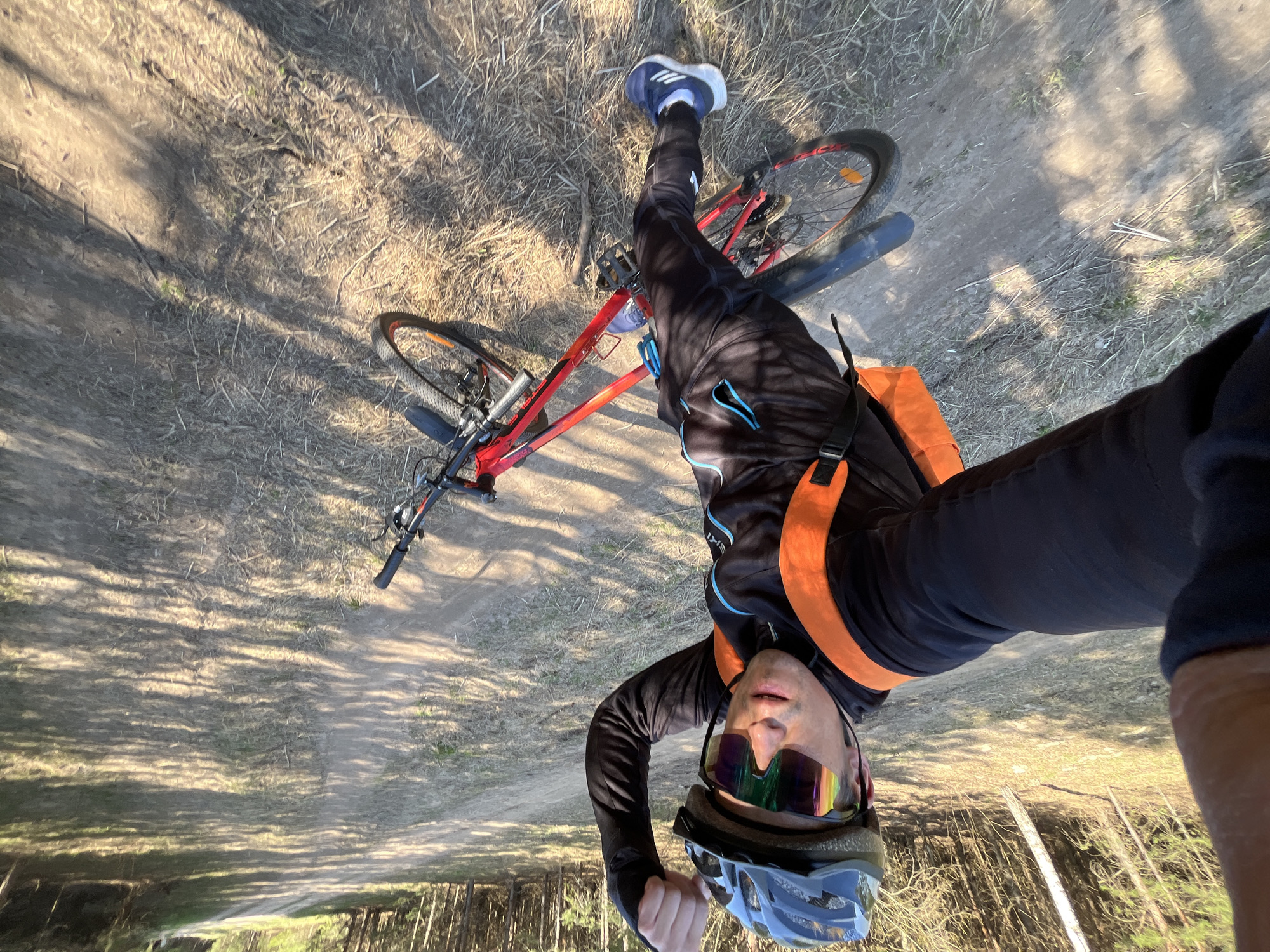 A cyclist is taking a selfie in a forest environment. The cyclist is wearing a helmet and sunglasses, and a jacket. They are straddling a red bicycle. A path is visible through the trees. The location is likely in Chaikovsky, Russia.