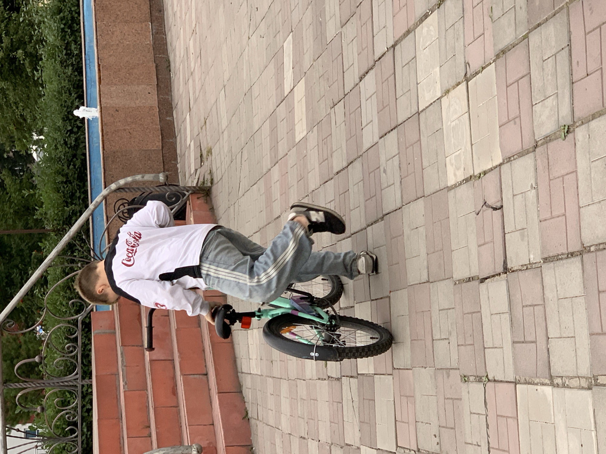 A boy, seen from behind, walks a turquoise bicycle on a brick-paved area. He wears a white long-sleeved shirt with "Coca-Cola" written on the back in red, and light blue pants with white stripes down the sides. The bicycle is small with a black seat and handlebars. Behind him, red brick stairs ascend to an ornate metal railing and lush green bushes. To the right, a fountain sprays water into a blue-lined basin, set against a textured brown wall. The overall scene appears to be an outdoor public space, possibly a park or plaza, in Prokopyevsk, Russia.