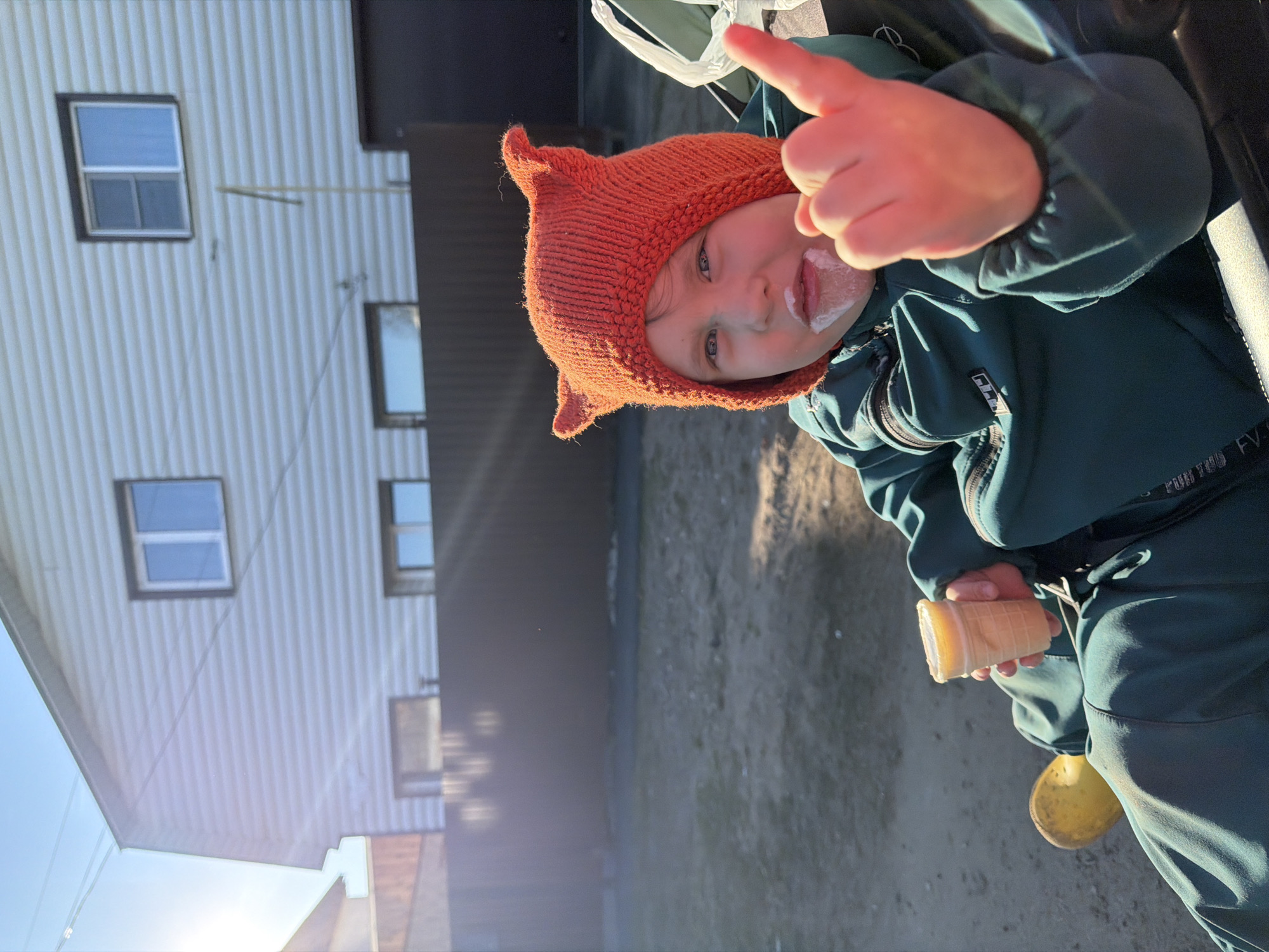 A young child, likely a toddler, is the focal point of the image. The child has light skin and blue eyes, and is wearing a bright orange knitted hat with small ears on top. There is ice cream smeared on the child's face. The child is wearing a dark green jacket and is holding an ice cream cone in one hand while pointing a finger with the other.
The background reveals a building with light-colored siding and dark trim. Windows with dark frames are visible on the building. The ground appears to be a dirt or sandy surface.
The location is identified as Chaikovsky, Russia, based on provided information.