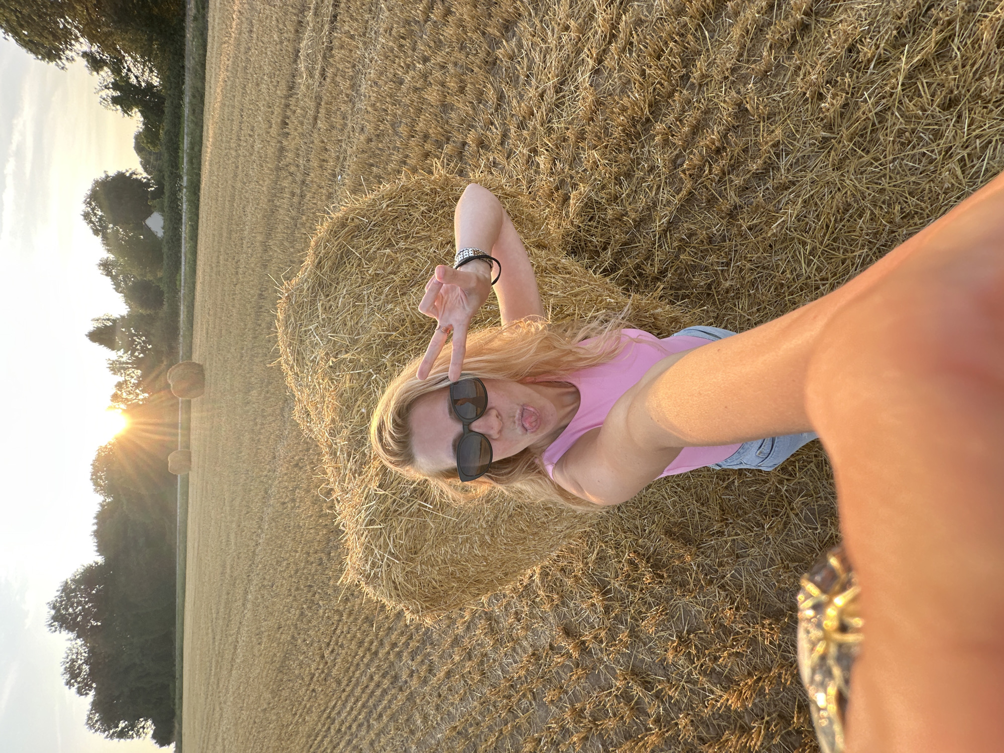 In a rural field in Trouville-Alliquerville, France, a blonde woman with blonde hair, wearing sunglasses, a pink sleeveless top, and blue jeans, is taking a selfie. She is sticking her tongue out and making a peace sign with her right hand. She has a bracelet on her left wrist. In the background, two hay bales are visible. The field is covered in straw. The sun is setting in the background, casting a warm glow over the scene.