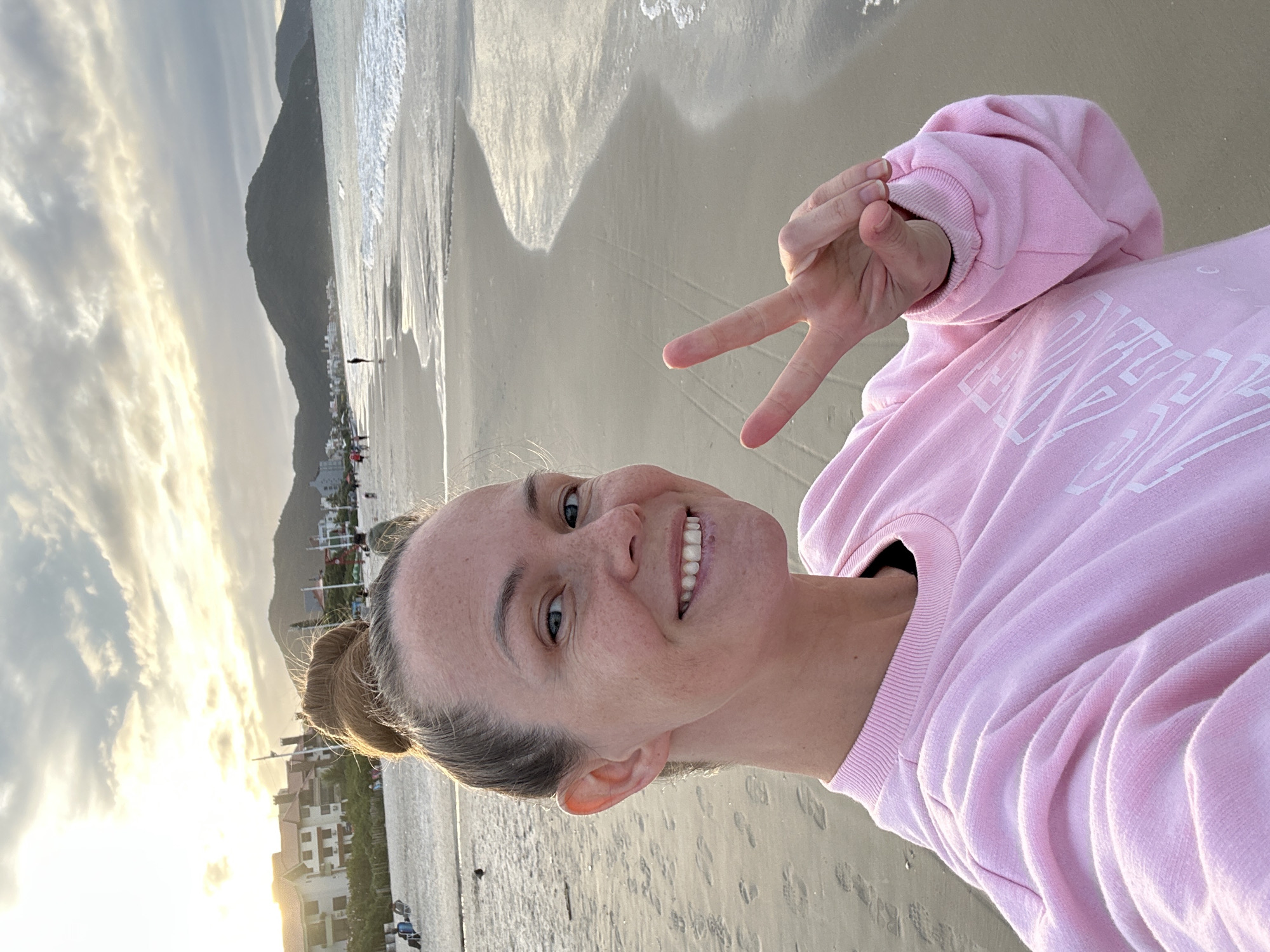 A woman takes a selfie on a beach in Florianópolis, Brazil. She is smiling, holding up a peace sign, and wearing a pink sweatshirt. The beach has light-colored sand and a few buildings can be seen in the background. The water is to her left, and a mountain is to the far left. The sky is cloudy and the sun is beginning to set.