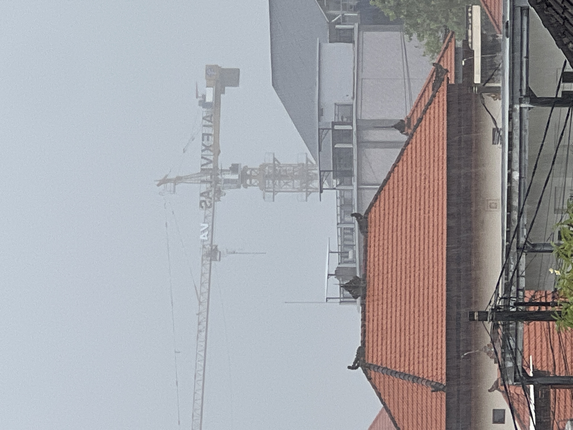 The image captures an outdoor scene dominated by a construction site under heavy mist or fog. In the foreground on the right, a prominent building features a reddish-orange tiled gabled roof. Portions of other buildings and structures are visible behind and to the left of this roof, extending into the midground. To the far right edge of the frame, power lines or cables are strung across utility poles, with some green foliage from a tree visible at the top right.
The central and left portion of the midground and background is occupied by a large, yellow construction tower crane. Its long jib extends horizontally across the misty sky. On the vertical mast of the crane, partially obscured by the atmospheric conditions, the text "ALEXIA" is visible, appearing reversed as "AS A I X E L A" when read from top to bottom on the crane's structure. Beyond the crane's mast, another structure, possibly a building under active construction or recently completed, is discernible through the heavy mist.
The prevailing weather condition is thick mist or fog, which significantly reduces visibility across the scene and creates a soft, diffuse light. The sky is a uniform light grey, indicating heavily overcast conditions. The overall impression is that of a bustling construction environment in North Kuta, Indonesia, experiencing a heavily overcast and foggy day. The time of day is not precisely determinable due to the atmospheric conditions, but the ambient light suggests it is daytime.