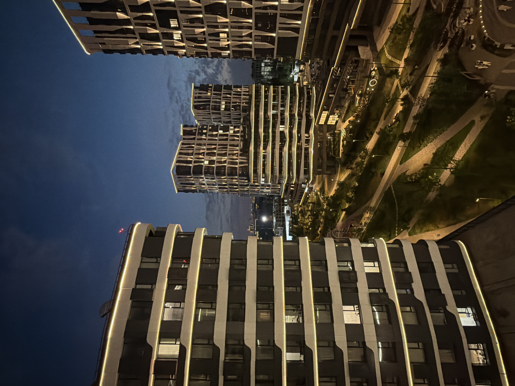 The image depicts a modern urban landscape at twilight, likely in Moscow, Russia. The foreground features the side of a multi-story building with a dark facade and prominent horizontal bands of warm yellow lighting accentuating its curved structure and balconies. Across a landscaped area, several other contemporary high-rise buildings are illuminated, showcasing their unique architectural designs with patterned facades and vertical lighting elements. The landscape between the buildings includes winding pathways, green spaces, streetlights, and subtle playground-like structures, all softly lit, creating an inviting atmosphere. The sky above is a deep blue with scattered clouds, indicating the transition from day to night. There are no visible people or ongoing actions in the scene, presenting a still, architectural study of a developed urban environment.