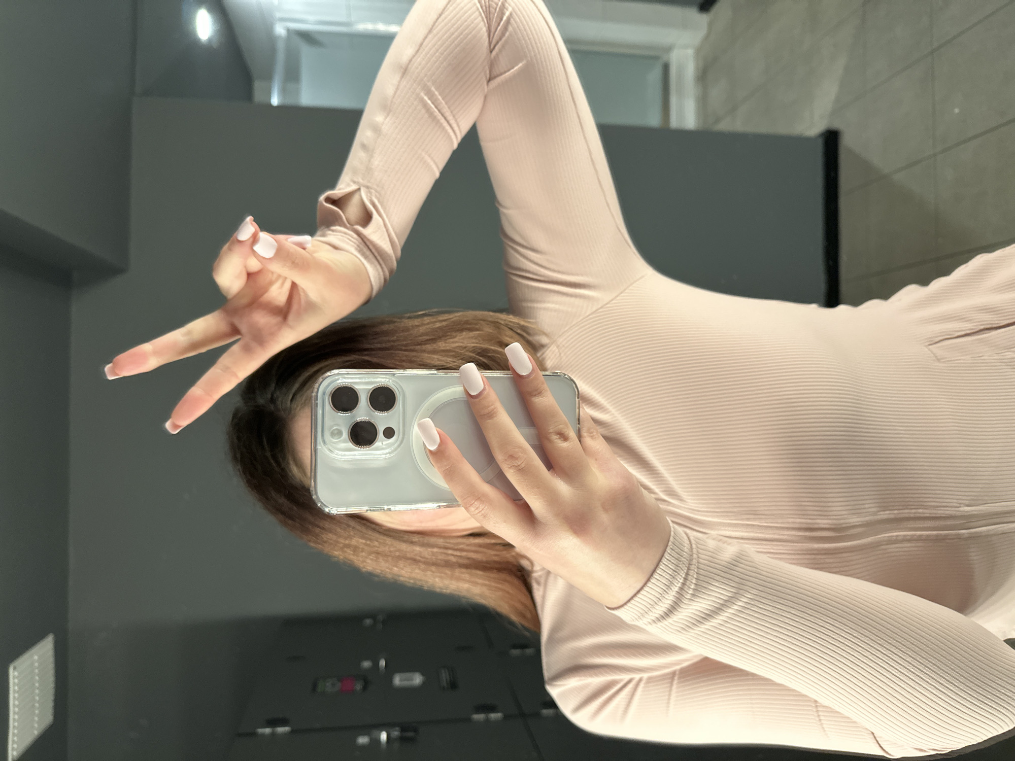 In a locker room environment, a person with brown hair is holding a smartphone in their right hand, taking a selfie in a mirror. Their left hand is raised, with the index and middle fingers extended in a peace sign. The person is wearing a light pink, ribbed long-sleeved shirt with thumbholes. The background shows grey lockers and a tiled floor. The scene does not contain any identifiable landmarks or elements that would place it in Carlow, Ireland.