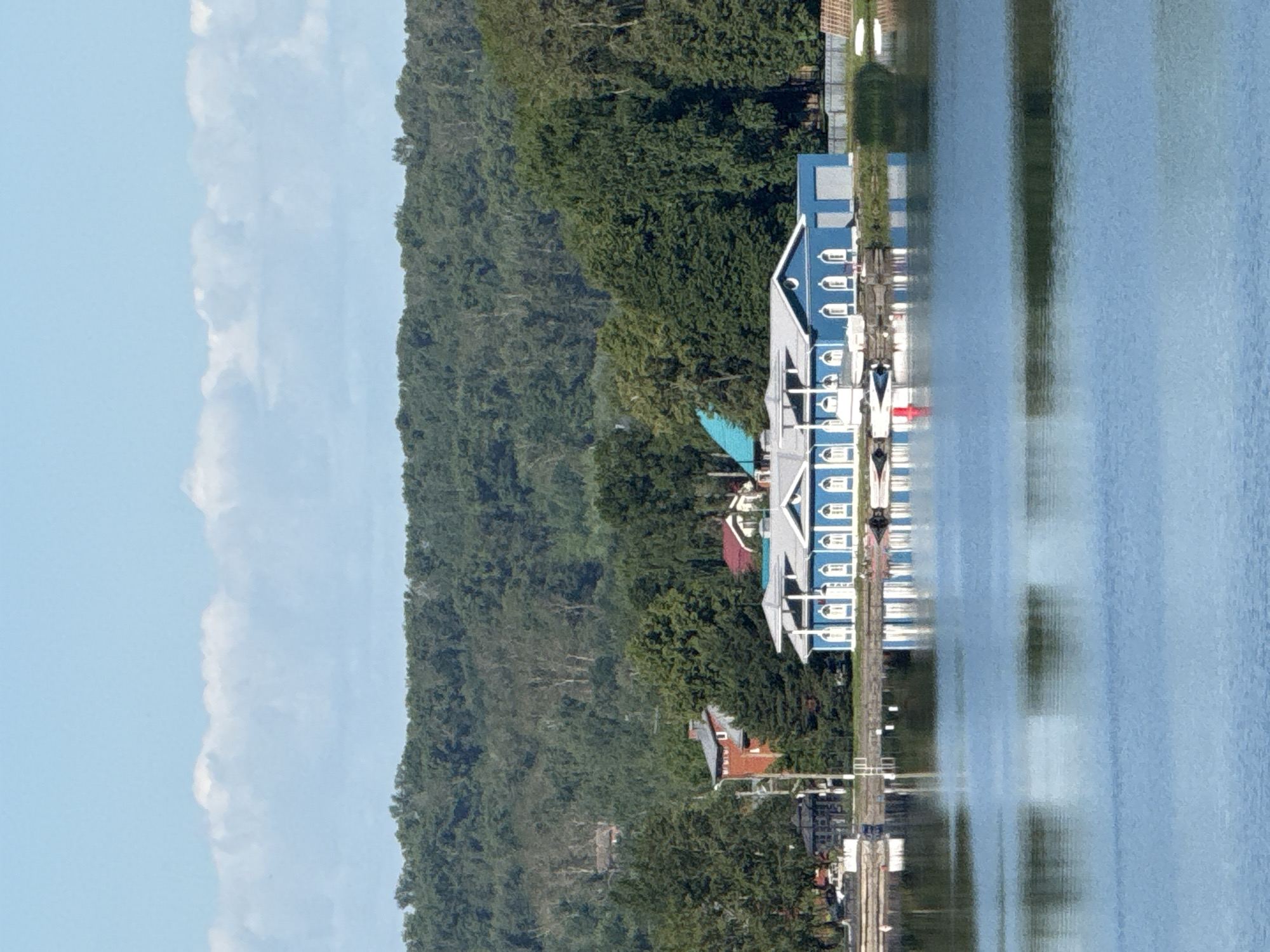 This is a horizontal, medium-distance, eye-level shot of a body of water with a blue building with white trim on the far bank, in Votkinsk, Russia. The water is calm and reflects the blue sky, the building, and the surrounding trees. The building is long and rectangular, with a white roof and many arched windows. In front of the building, there are several small boats. The far bank is covered in lush green trees, and behind them, a hill rises steeply, also covered in trees. The sky is a clear, light blue with a few wispy white clouds. The overall scene is peaceful and serene.
