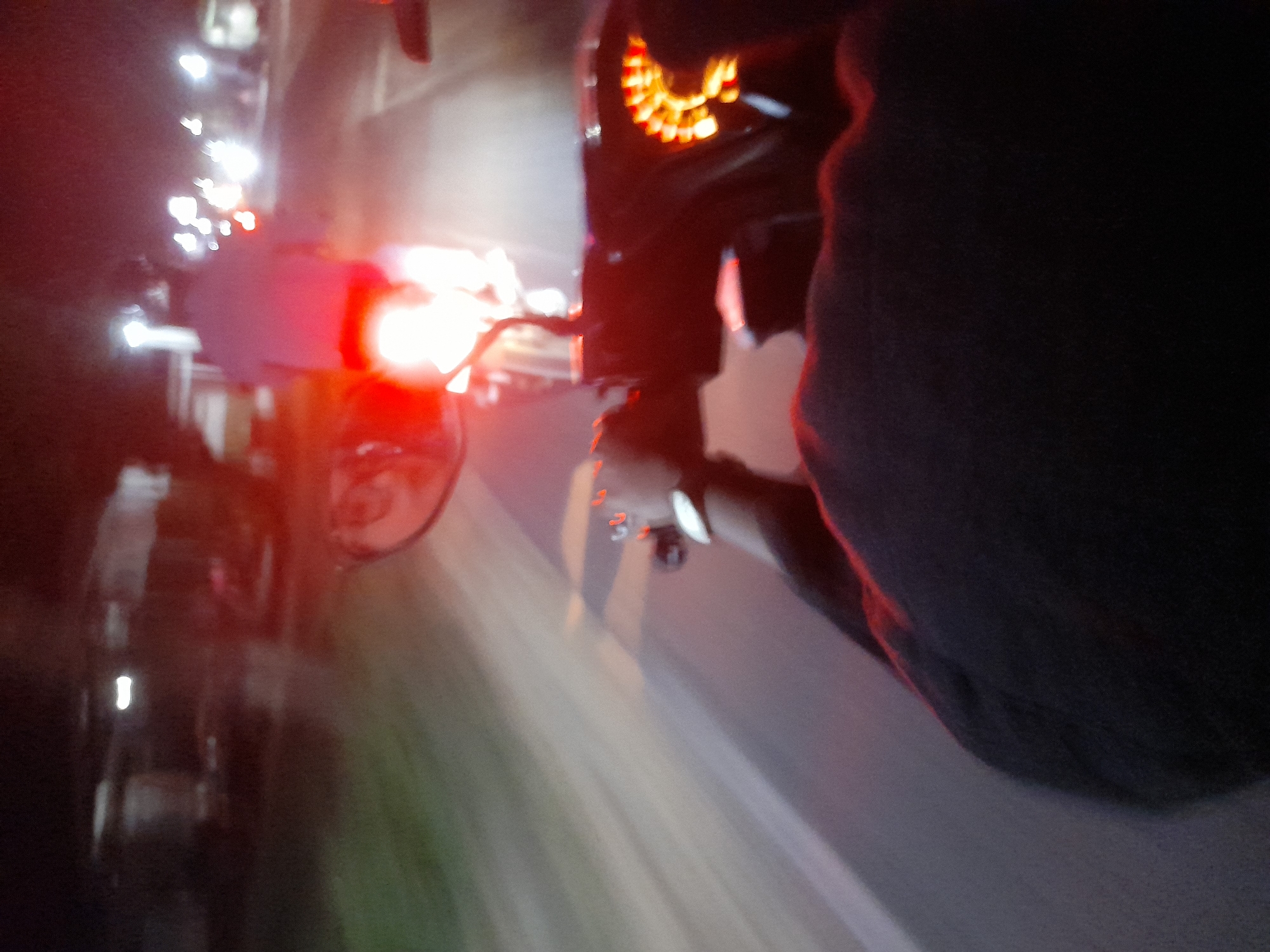 The image depicts a nighttime scene from the perspective of someone riding on the back of a motorcycle in Jetaksari, Indonesia. The motorcycle is moving, indicated by motion blur in the background and foreground. The rider in front is partially visible, wearing a dark shirt. The motorcycle's tail lights are illuminated, casting a red glow. Ahead, other vehicles and streetlights are visible, contributing to the overall blurriness of the scene. The road appears to be paved and there are indications of vegetation or structures along the side of the road. The lighting is low, characteristic of nighttime urban or suburban environments. The image captures a dynamic moment of travel.