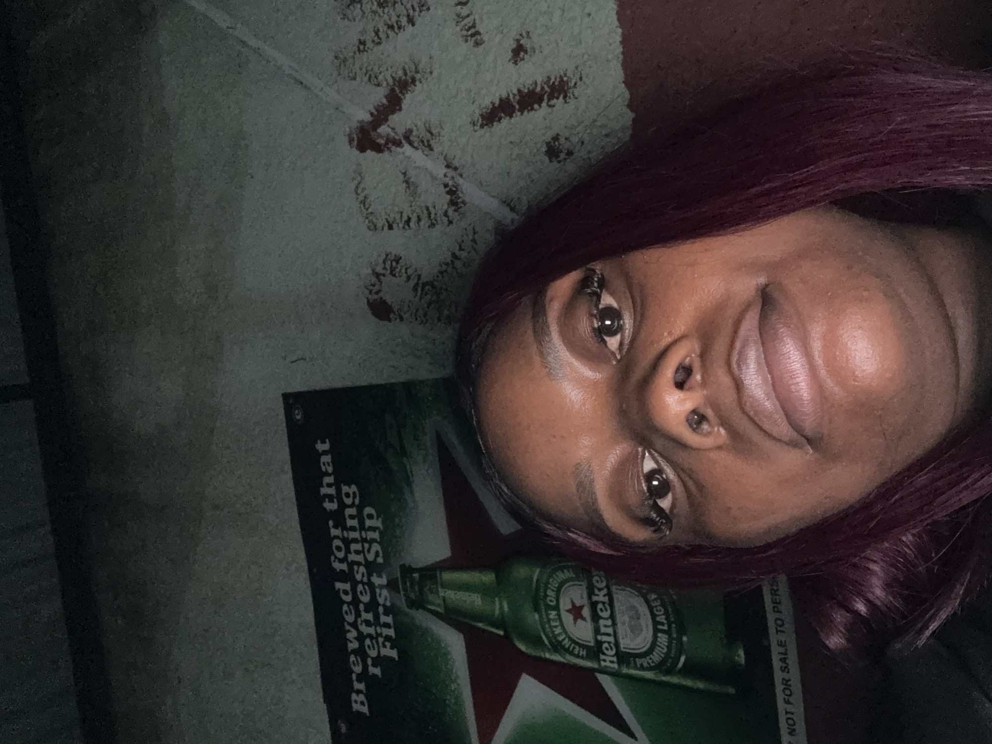 A close-up selfie is taken from a low angle of a young Black woman with purple hair and false eyelashes. She has dark skin, dark eyes, and full lips. She is looking up at the camera. Behind her is a poster advertising Heineken beer, showing a bottle of Heineken with the tagline "Brewed for that refreshing First Sip". The poster is green and red. The wall behind the poster appears to be concrete and has some writing on it, though it is difficult to read. The overall lighting suggests an indoor, possibly dimly lit environment. The image does not provide any specific clues about the location in Abuja, Nigeria.
