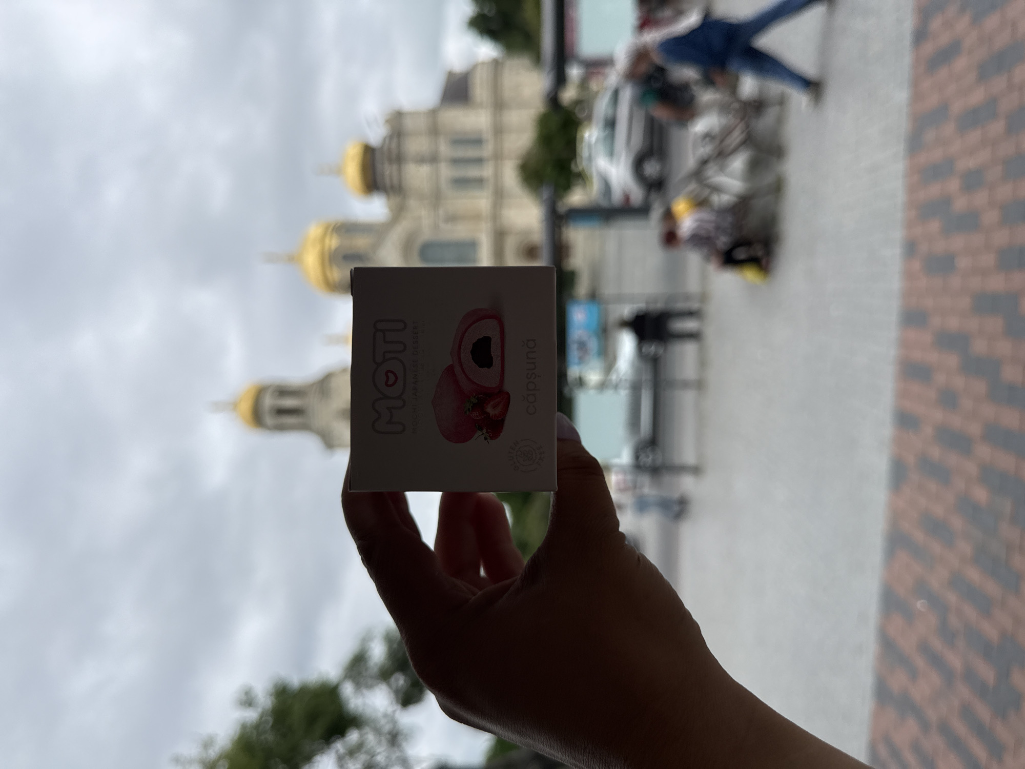 A person's hand holds a small, rectangular box of "MOTI MOCHI JAPANESE DESSERT" featuring a picture of a strawberry mochi and the word "căpșună". The box is positioned in the foreground. In the blurred background, a large building with golden domes, possibly a church or cathedral, is visible against a cloudy sky. Several indistinct figures of people and vehicles are also present in the outdoor urban environment.