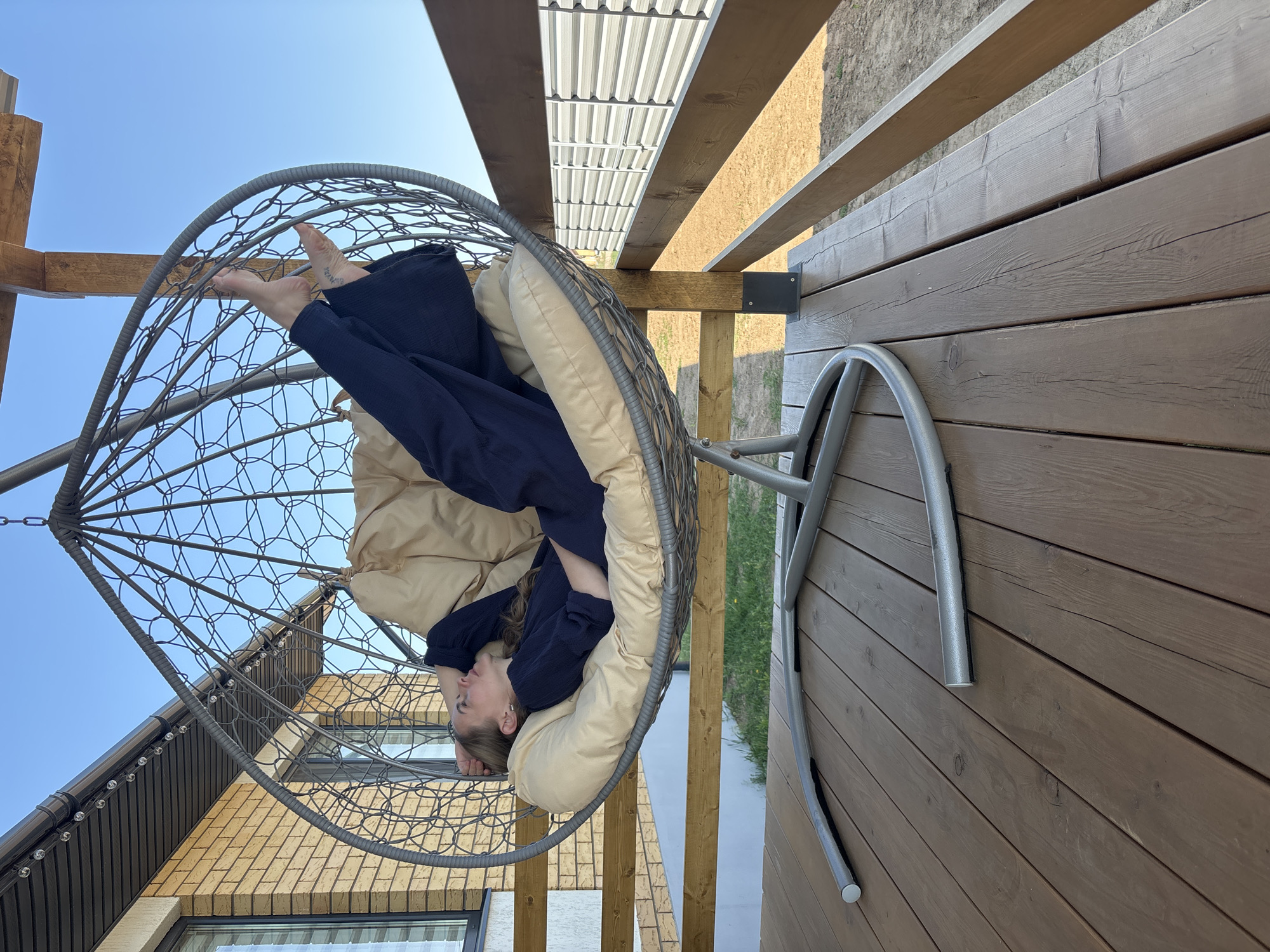 A woman rests in a hanging chair on a wooden deck. The chair, constructed of a grey metal frame with a woven pattern, cradles a beige cushion. The woman, wearing dark blue pants and a matching shirt, reclines with her legs elevated towards the top of the chair. Her head rests against the side, her eyes closed.
The deck is constructed of parallel wooden planks and is partially covered by a wooden pergola with a corrugated metal roof. Beyond the deck, glimpses of a lawn and possibly a body of water can be seen. A brick building is visible in the lower left corner.
The photograph was taken in the city of Chaikovsky, Russia.