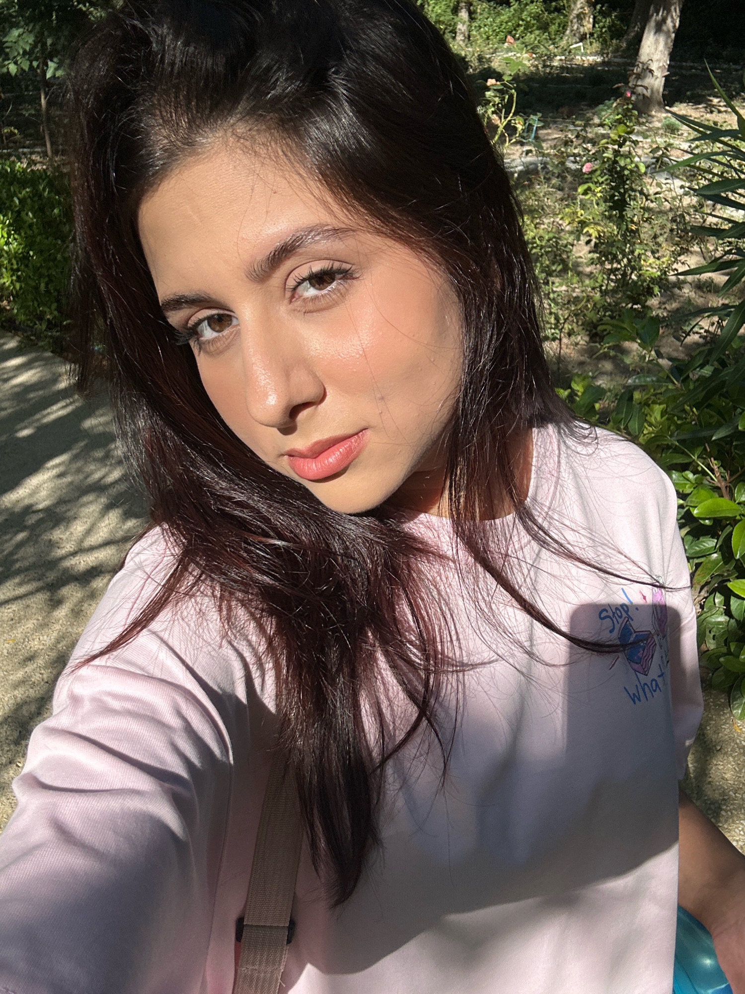 A young woman with dark hair and brown eyes is captured in a selfie taken outdoors. She is wearing a light pink t-shirt with a graphic design that includes text and a butterfly. The background suggests a garden or park setting, with trees, foliage, and dappled sunlight. The woman's face is turned slightly towards the camera, and she has a neutral expression. The image appears to be taken during the daytime, with natural lighting. The location is specified as Sabail Raion, Azerbaijan.