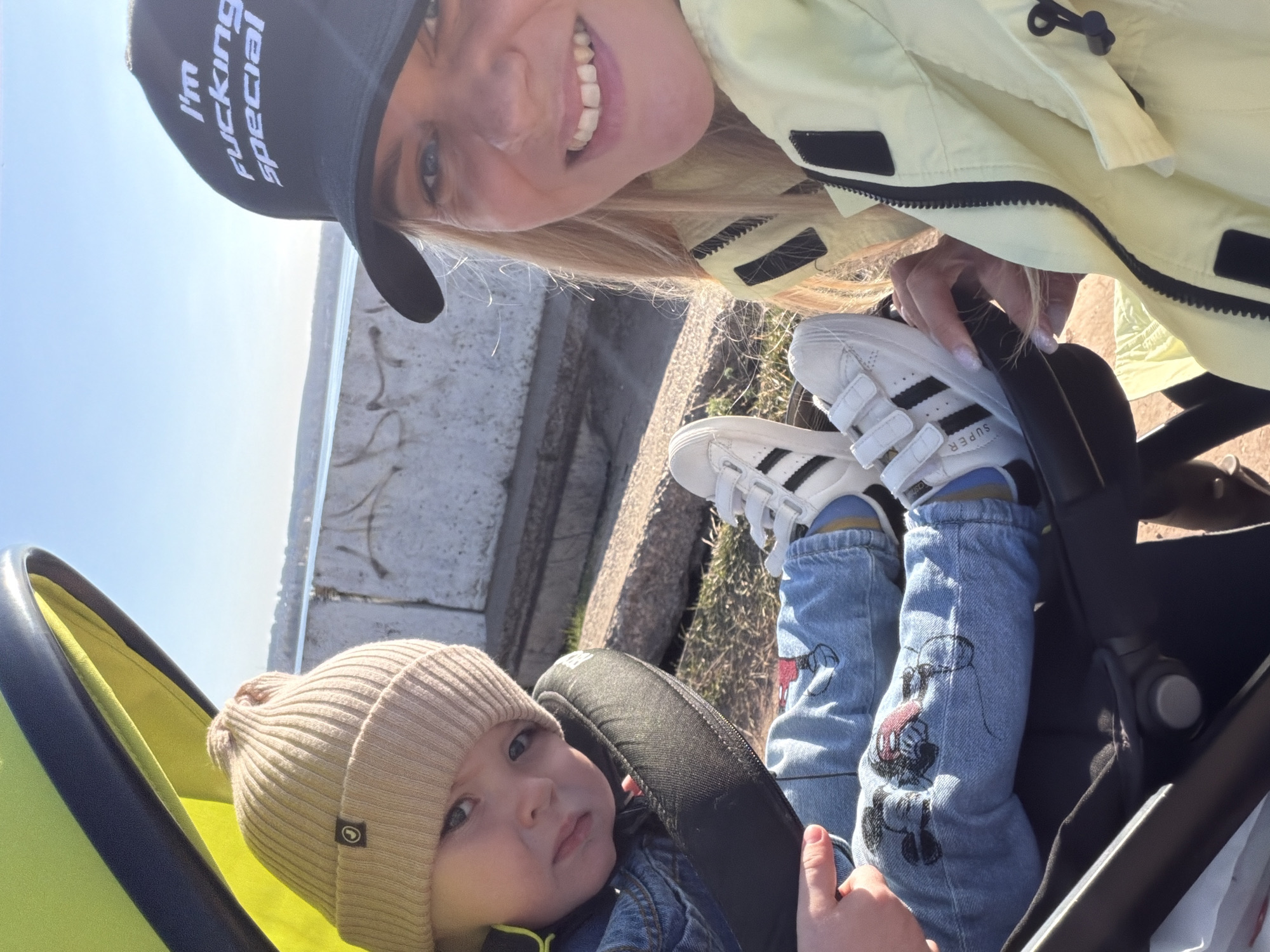 A medium shot captures a fair-skinned woman and child in Votkinsk, Russia. The woman, wearing a black baseball cap with text, and a light yellow jacket, holds a phone at a high angle, smiling. In front of her, a young child in a tan beanie, denim jacket, and Mickey Mouse-print jeans sits in a stroller. The child wears white Adidas sneakers with black stripes. The background consists of a concrete structure with sunlight reflecting off the adjacent water.
