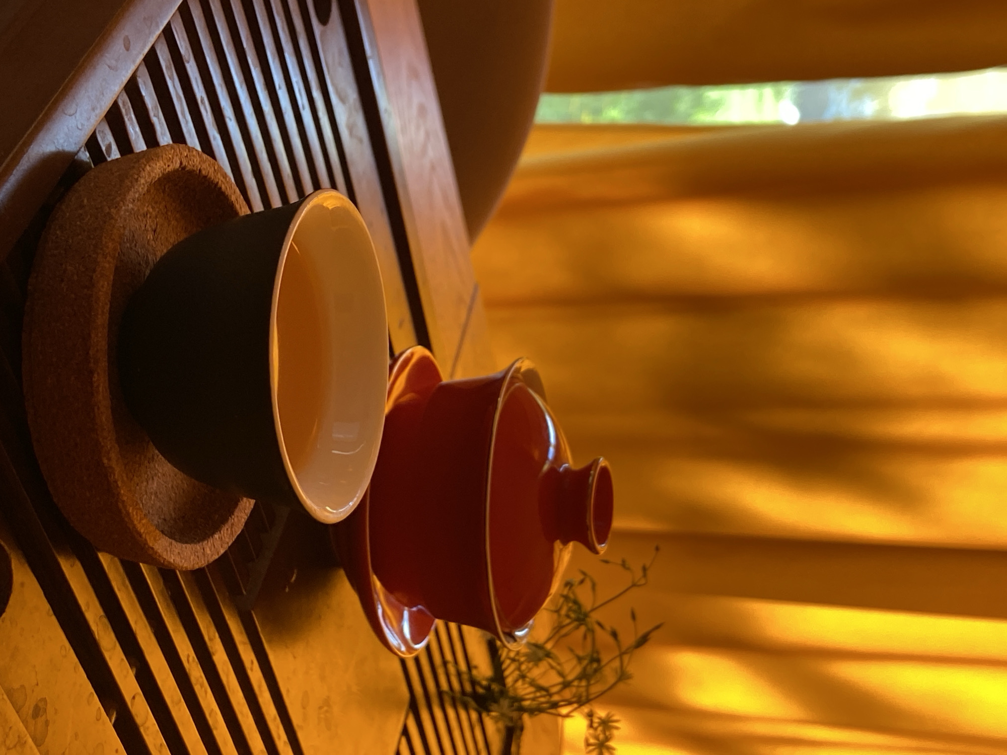 A close-up shot depicts a tea set arranged on a wooden surface, likely a tea tray, with sunlight filtering through a window with orange curtains in the background. A dark-colored tea cup sits within a cork coaster on the left. To the right, a red lidded tea bowl rests on a saucer; dried floral sprigs are near the red tea bowl. This scene might be indoors in Tyumen, Russia.