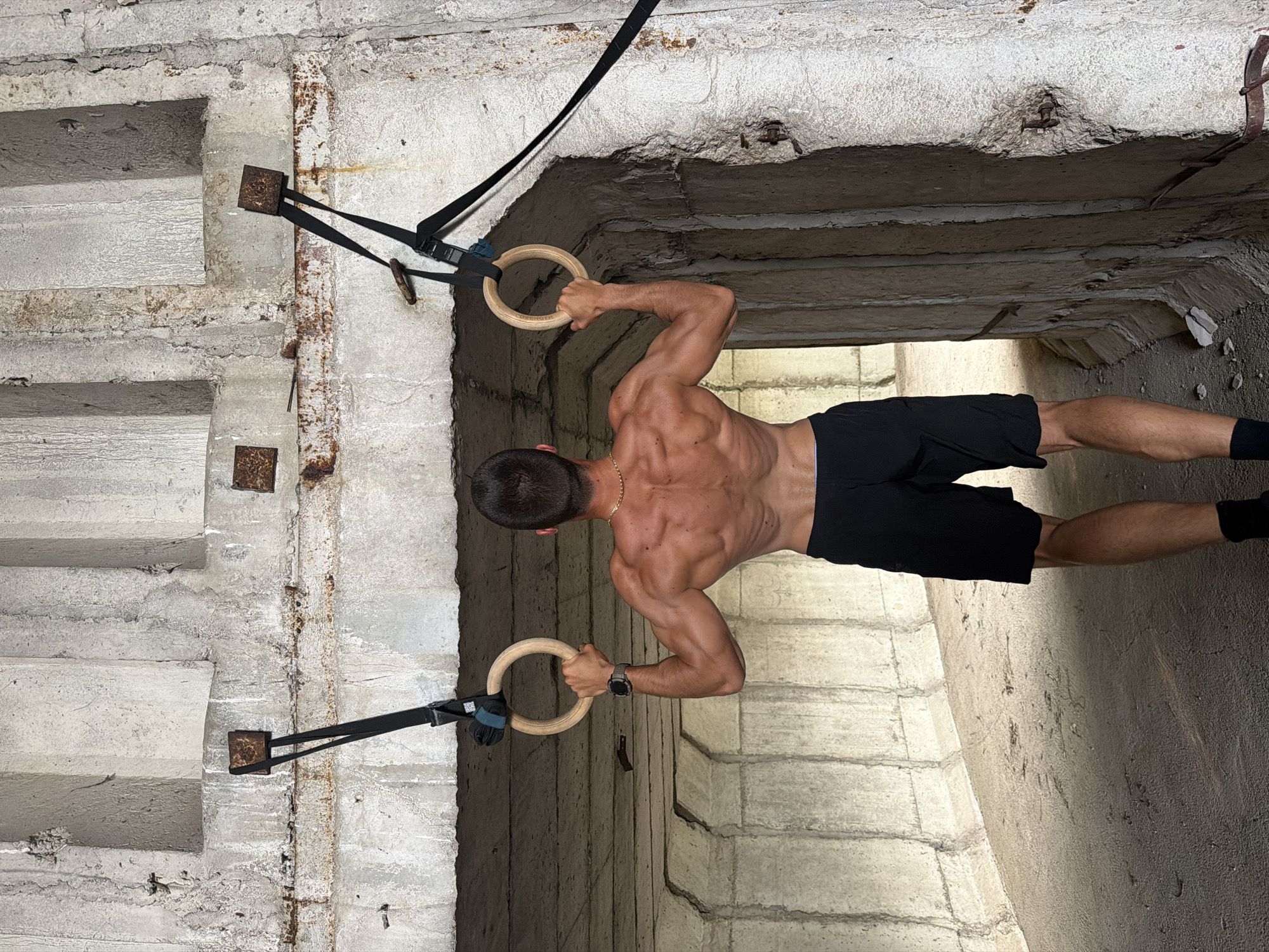 A male subject is depicted from the rear, shirtless, wearing black athletic shorts and black socks, positioned within an austere concrete structure in Bogdanitsa, Bulgaria. He has short dark hair, a gold neck chain, and a dark-colored smartwatch on his left wrist. The subject exhibits a developed muscular physique, particularly in the back and arms. He is actively engaged in a calisthenics exercise, gripping two wooden gymnastic rings with both hands. The rings are suspended by black nylon straps, which are affixed to rusted square metal plates embedded into the concrete overhead structure.
The environment consists of a large, weathered concrete tunnel or underpass. The concrete surfaces display significant age, characterized by discoloration, surface deterioration, and extensive rust staining originating from various embedded metal components. The interior walls and ceiling feature a segmented construction with prominent vertical and horizontal seams. Light enters the structure from both the foreground and background, illuminating the scene. The ground surface appears to be coarse earth or deteriorated concrete.