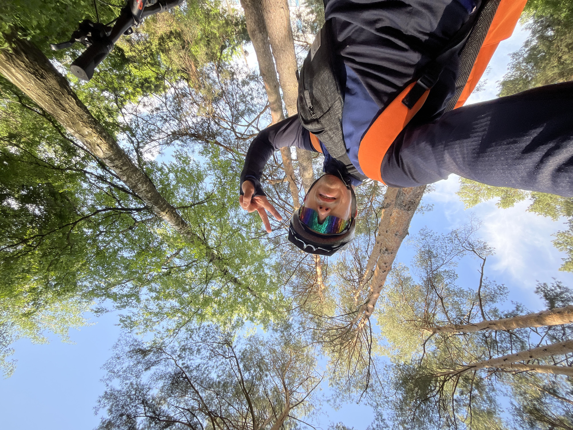 The image is a low-angle selfie taken in a forest in Chaikovsky, Russia. A person wearing a cycling helmet with rainbow-colored lenses and a dark blue and orange athletic jacket is looking down at the camera and making a peace sign. They are wearing a dark backpack. Above the person are trees with green and gray foliage against a blue sky. The handlebars of a bicycle are visible in the upper left corner. The camera is angled upwards towards the canopy.