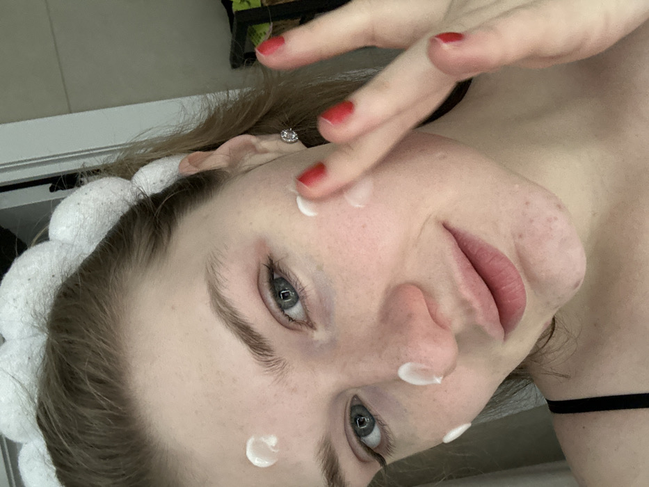 A young woman applies white cream to her face. She wears a white, puffy headband and has red nail polish. Her skin is fair, and she has visible pores. The image is taken at a close-up angle, showing her face and a hand with red nails. Her expression is relaxed and slightly smiling. A thin black strap is visible around her neck, suggesting clothing. Her eyes are light blue.