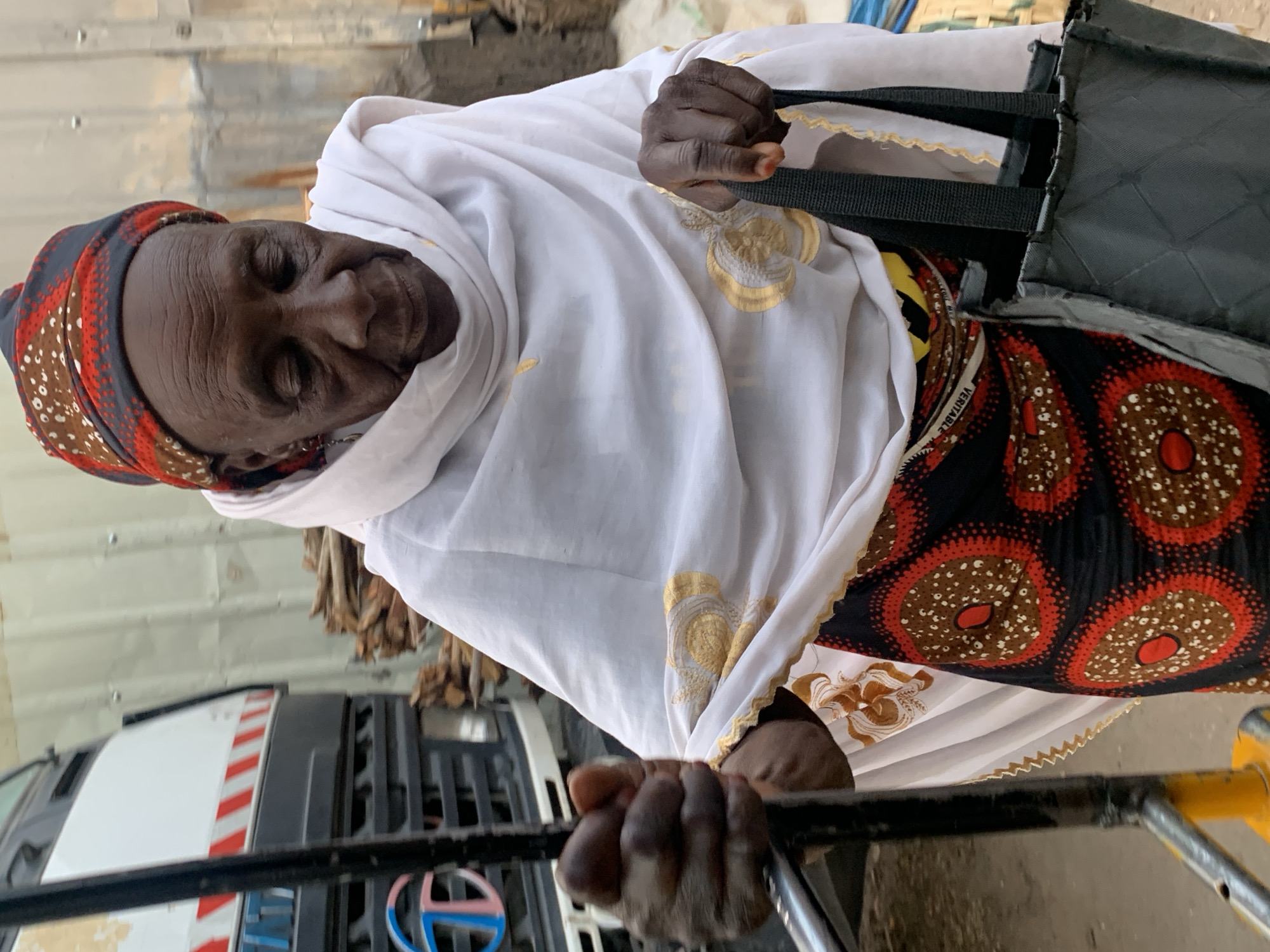 In Jos, Nigeria, a dark-skinned elderly woman wearing a red and brown patterned headscarf and a white shawl with gold embroidery holds the handlebar of a rolling cart. Her dress is printed with large brown and red circles on a black background. The woman is holding a dark grey bag with black straps. The background includes a stack of firewood and a white and grey truck.