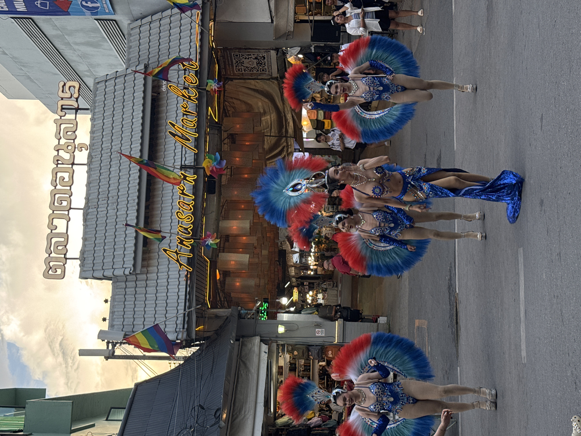 The image depicts a street scene in Chiang Mai City Municipality, Thailand. In the foreground, four performers in blue, red, and white Carnival-style costumes are visible on the road. They are standing and posing. Behind them is a building with a tiled roof and a sign that reads "Anusarn Market" in English and Thai. Rainbow flags are displayed above the market entrance. The street appears busy, with people and other objects visible in the background.