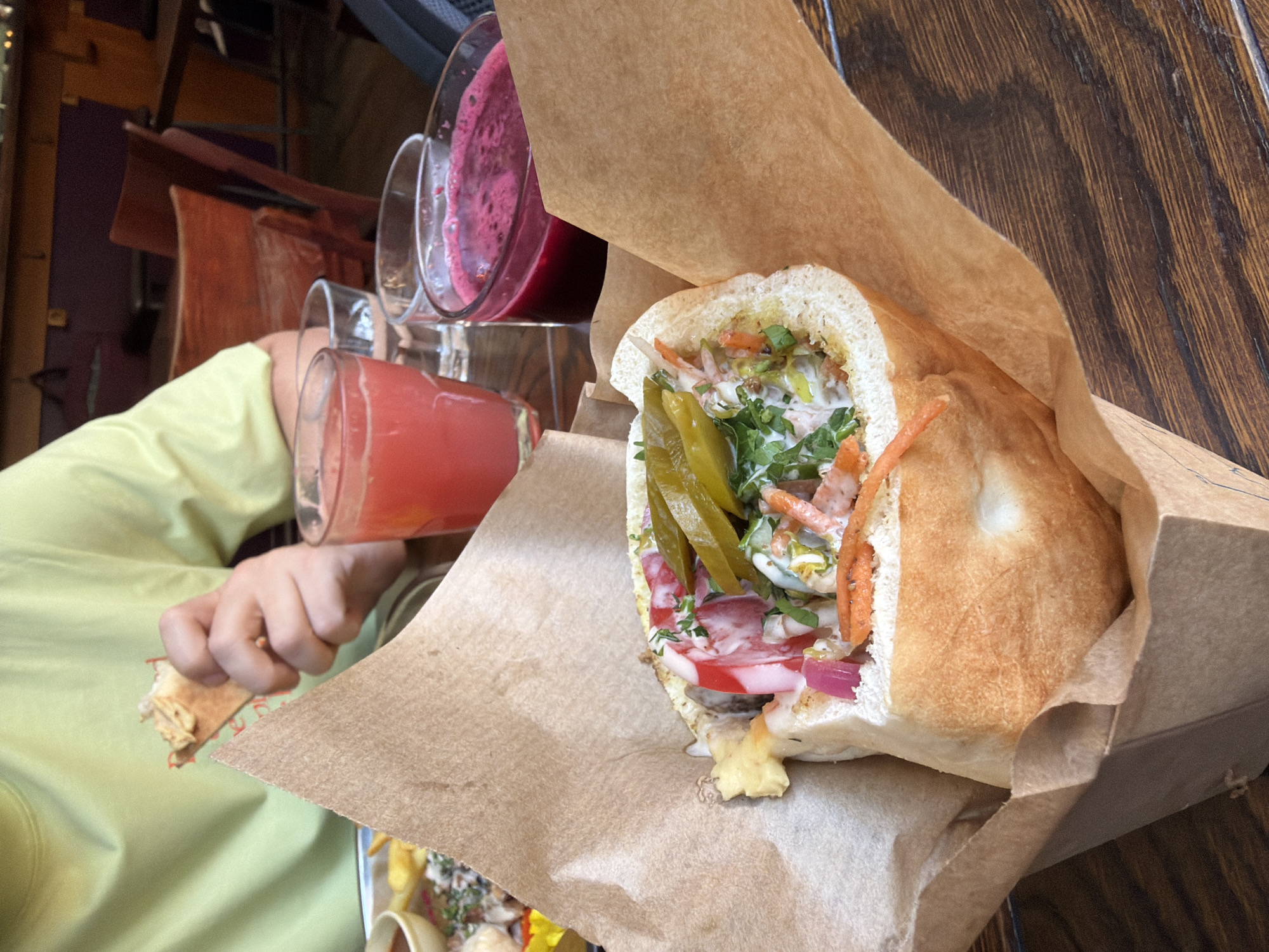 The image captures a casual dining scene within a restaurant, presumed to be located in Saint Petersburg, Russia. In the immediate foreground, a large, oblong sandwich, appearing to be a filled flatbread or a short baguette, is presented upright in a folded brown paper wrapper or container. The sandwich is generously filled with vibrant ingredients, including sliced red onions, shredded orange carrots, chopped green herbs (likely parsley or dill), a creamy white sauce, and prominent bright green pickled peppers on top. Some red elements, possibly tomato slices or a red sauce, are also visible within the filling, alongside a small yellowish piece of food near the left edge.
To the upper left, a person wearing a light green long-sleeved shirt is visible, holding a small piece of bread or pita, smeared with a light brown spread, in their right hand, suggesting they are actively eating.
On the dark brown wooden table, positioned to the right of the main sandwich, are three glasses. The leftmost glass contains a bright pink or light red juice, the middle glass holds clear water, and the rightmost glass contains a darker, deep purple or rich red juice with a slight foamy top.
The background shows an out-of-focus restaurant interior featuring wooden tables and chairs, some appearing reddish-brown. The walls are painted a deep purple or plum color, with hints of yellow or orange accents visible. A silver tray with other food items, including French fries and a small brown bowl, is partially visible behind the drinks and the person.