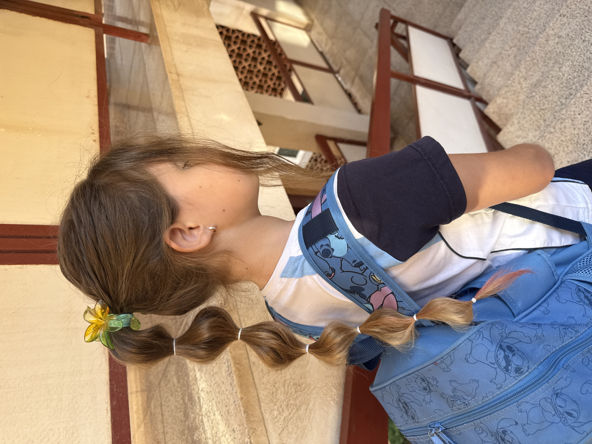 The image captures a young person from a side-back angle, facing towards the right side of the frame with their head tilted slightly upwards. They have long, light brown hair styled in a unique ponytail. The ponytail is gathered at the back of the head with a yellow and green flower-shaped hair clip and is then segmented into several sections using small white elastic bands, creating a series of wavy, bubble-like segments. The very end of the ponytail shows a faint reddish tint. A small, shiny earring is visible in their left ear.
The person is wearing a white t-shirt layered under a dark blue short-sleeved top or jacket. They are also carrying a light blue backpack adorned with outlines of the Disney character Stitch.
The background depicts an architectural setting, likely an outdoor or semi-outdoor corridor or courtyard. Features include light-colored walls, some with visible reddish-brown frames or beams, and a section with decorative terracotta-colored lattice work. In the distance, a set of concrete stairs leads upwards. The bright lighting suggests it is daytime, possibly sunny. There is no discernible text visible in the image. The person appears to be standing or moving through this architectural space.
