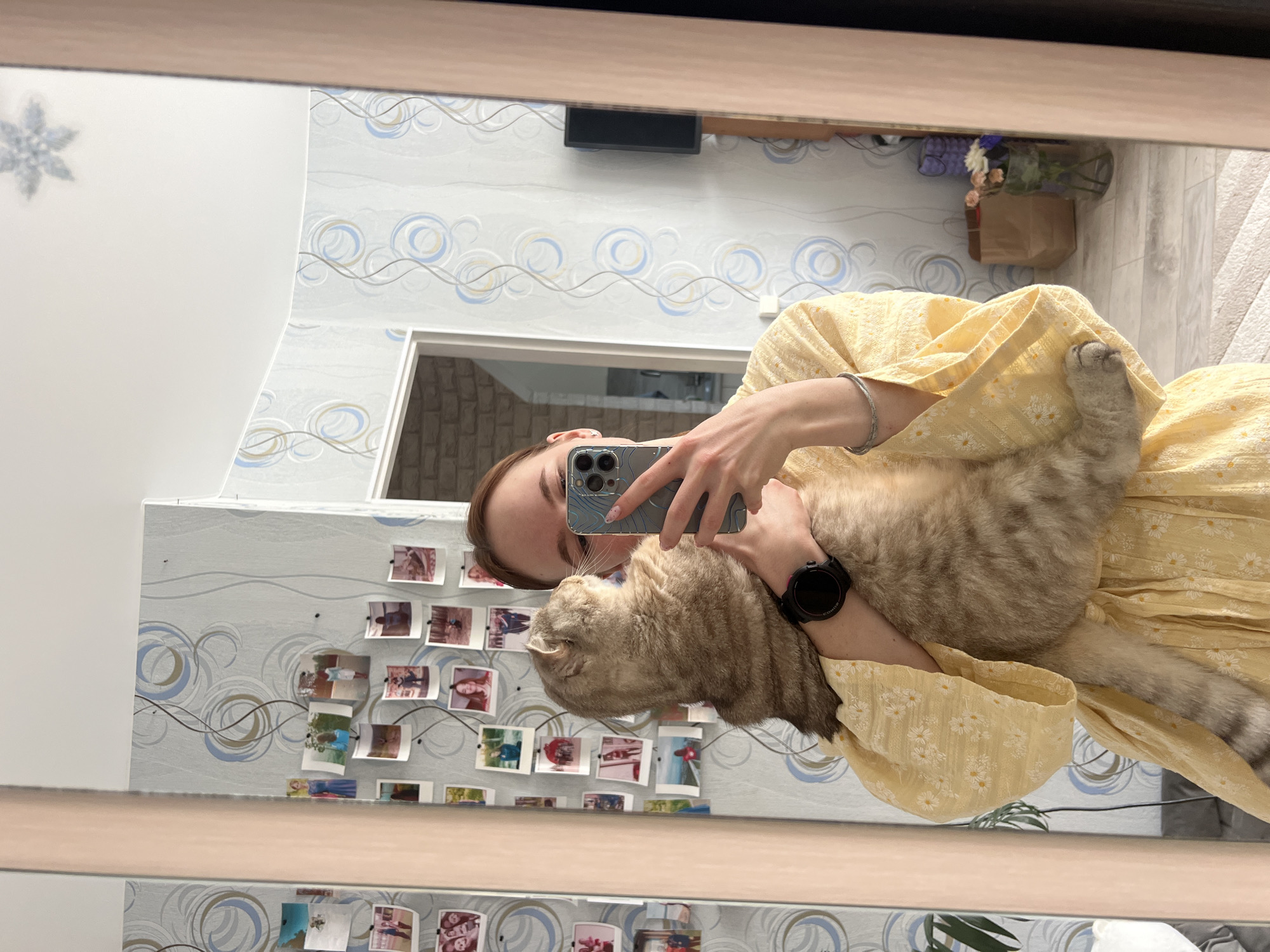 A mirrored image captures a woman holding a cat while taking a selfie with her phone. The reflection indicates the setting is likely indoors, featuring patterned wallpaper adorned with numerous photographs. Other details visible in the reflection include a doorway, a floral arrangement in a vase, and part of the room’s interior design. Based on geolocation research, this image was taken in Chaikovsky, Russia.