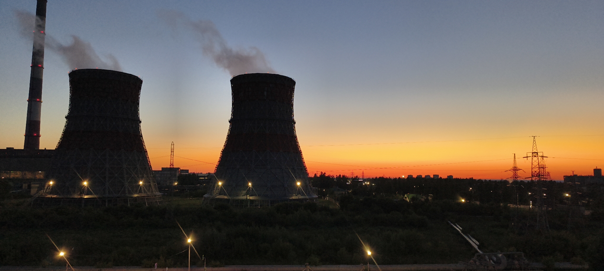 A wide-angle, outdoor, daytime shot captures two large cooling towers of a power plant at dusk. The towers are cylindrical with a wide base and taper towards the top, featuring red and grey bands and illuminated lights at their bases. Steam billows from the top of the tower on the left, while the tower on the right also emits steam. A tall, striped smokestack is visible to the left of the cooling towers, also emitting smoke. The foreground is dominated by dark green foliage and trees, with several illuminated lampposts. In the background, a low-lying cityscape with buildings and power lines stretches across the horizon. The sky transitions from a pale blue at the top to a vibrant orange and yellow near the horizon, indicative of sunset. The scene is set in an industrial area of an unknown city.