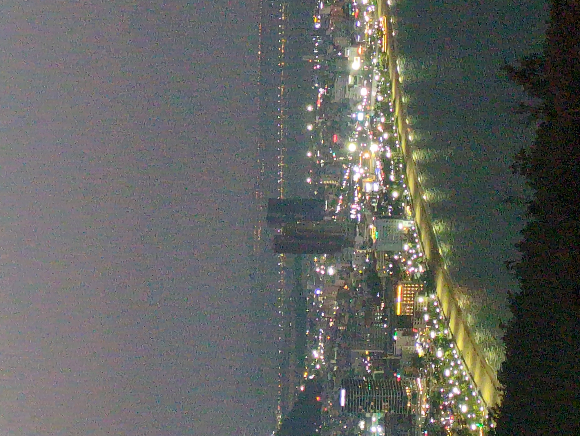 The image shows a night view of South Quy Nhon Ward, Vietnam. The cityscape is illuminated with numerous lights, contrasting with the darker areas of the sky and possibly a body of water on the right side of the frame. A line of lights could indicate a road or a coastal area. There are several tall buildings visible, and the general atmosphere is that of an active urban environment at night.